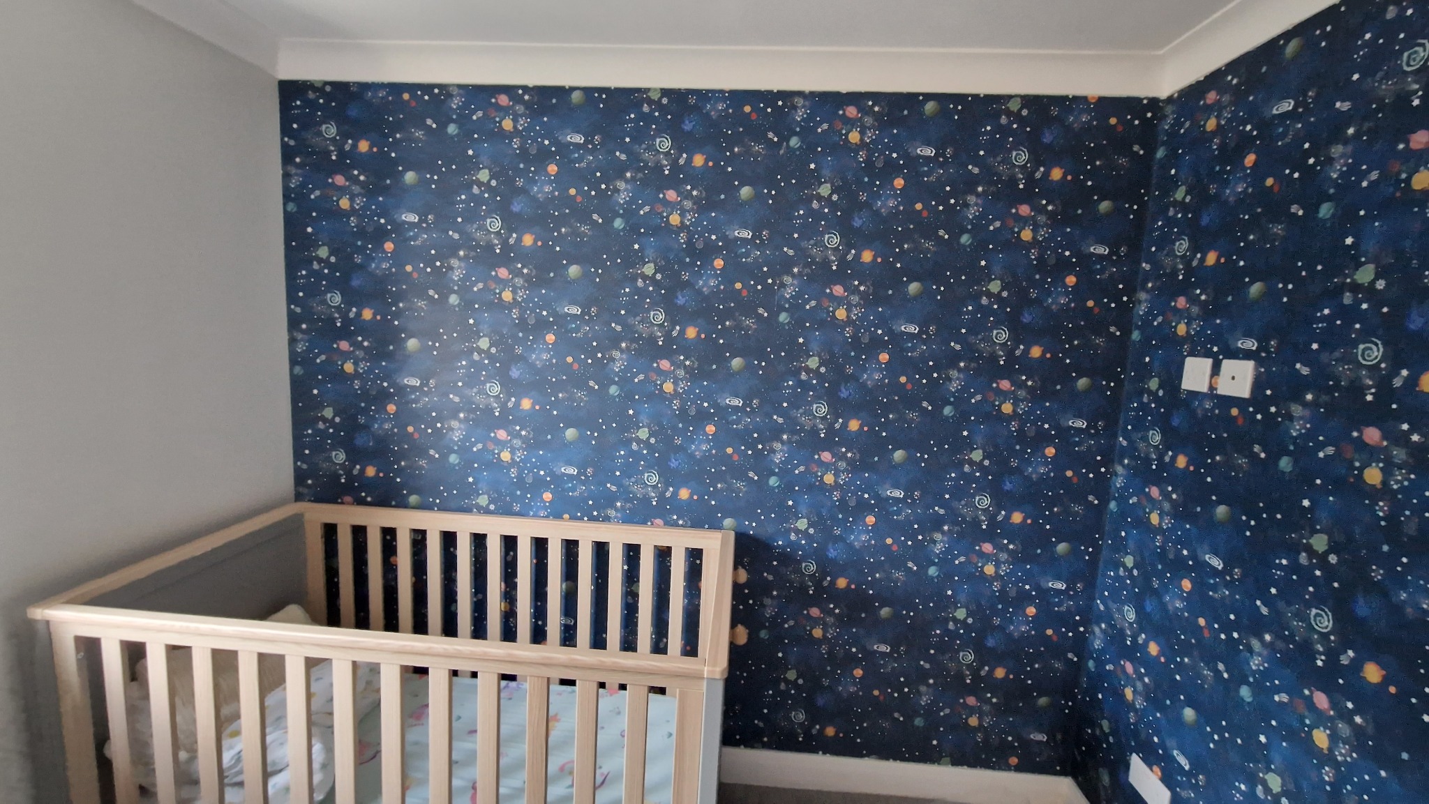 Pokój dziecięcy po remoncie: ściany pokryte tapetą w kosmiczne wzory, widoczne łóżeczko dziecięce. Wykończenie wnętrza z motywem galaktyki.