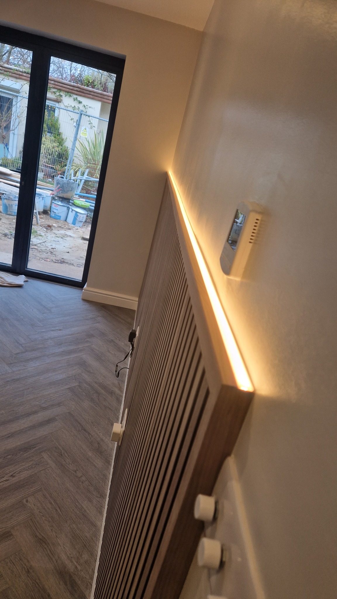 Nowoczesny panel ścienny z drewna z subtelnym oświetleniem LED, widoczne gładzie na ścianie i podłoga ułożona w jodełkę. Na zewnątrz okna widoczne prace budowlane.