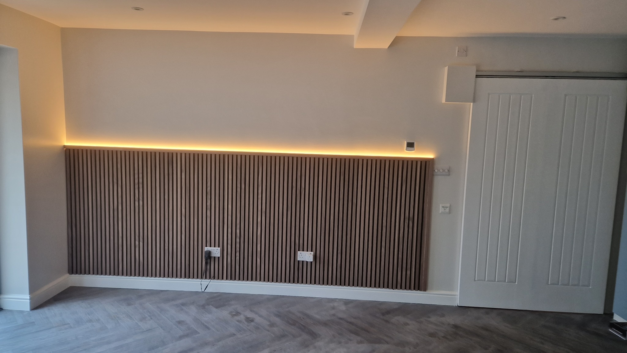 Panel lamelowy z podświetleniem LED na ścianie, widoczne gniazdka elektryczne i białe drzwi. Parkiet ułożony w jodełkę. Nowoczesne wykończenie wnętrza.