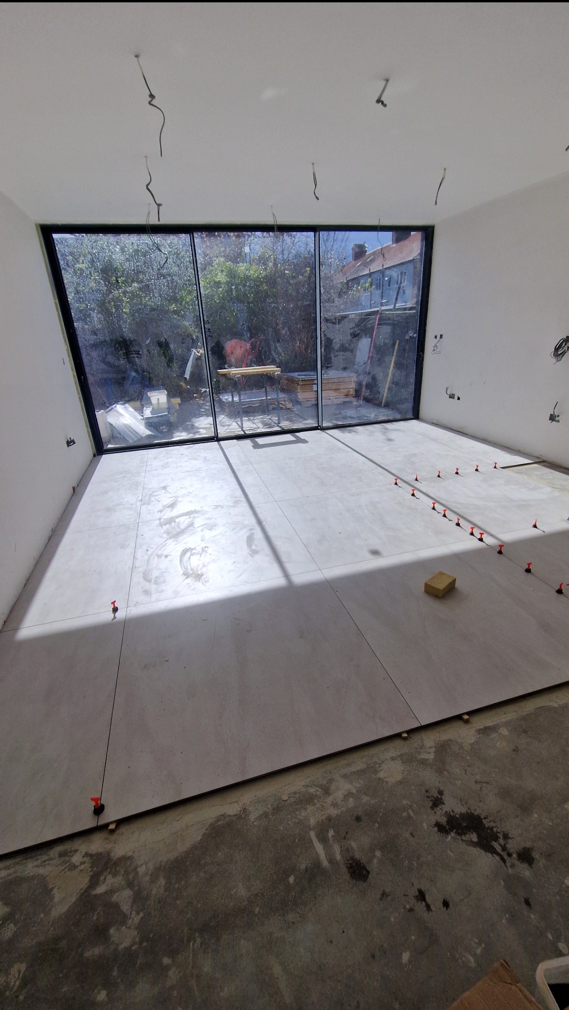 Jasne płytki podłogowe w trakcie układania, z systemem poziomowania, w przestronnym wnętrzu z dużym oknem na ogród. Widoczne niedokończone wykończenie podłogi.