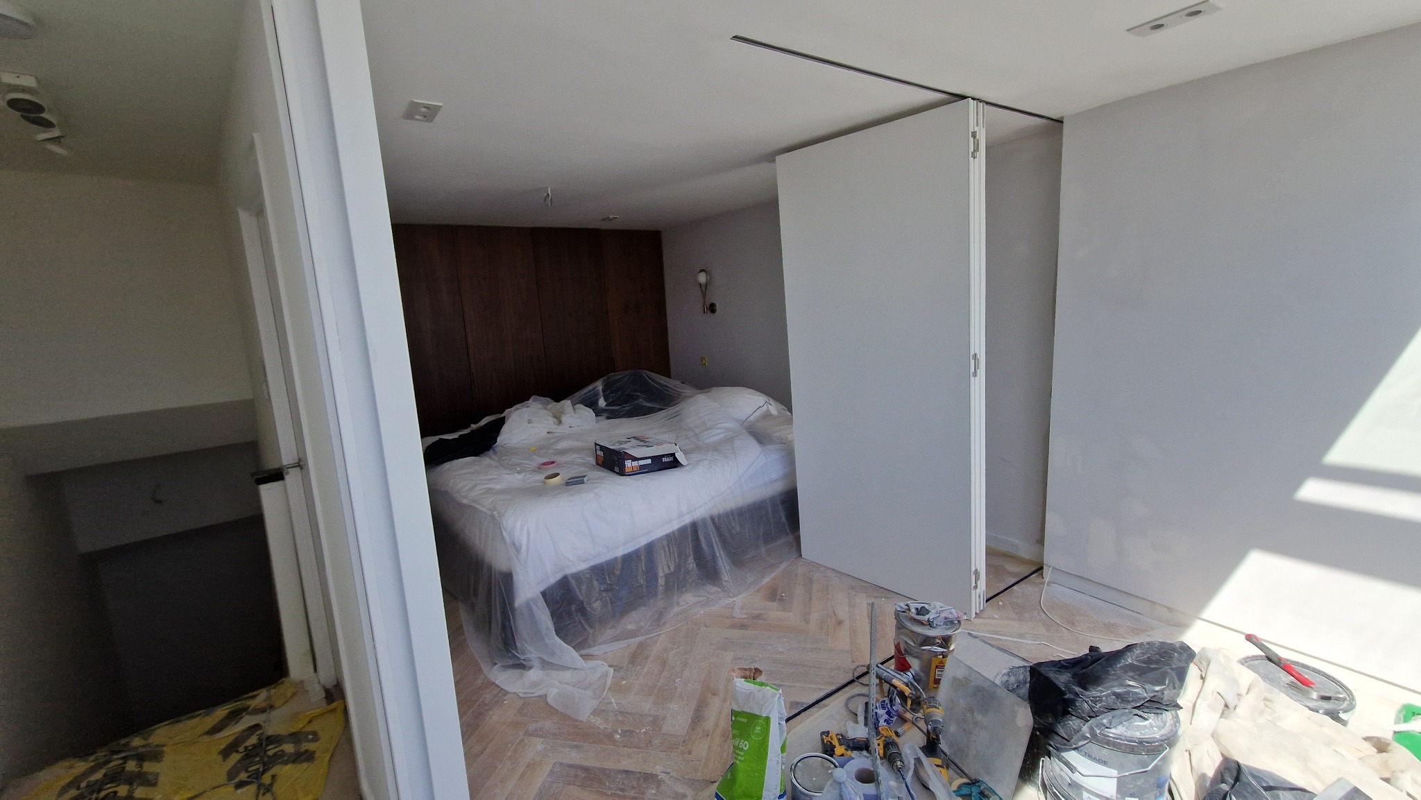 Pokój w trakcie malowania: zabezpieczone łóżko folią, widoczne narzędzia malarskie, świeżo pomalowane ściany na biało, drewniana podłoga w jodełkę.