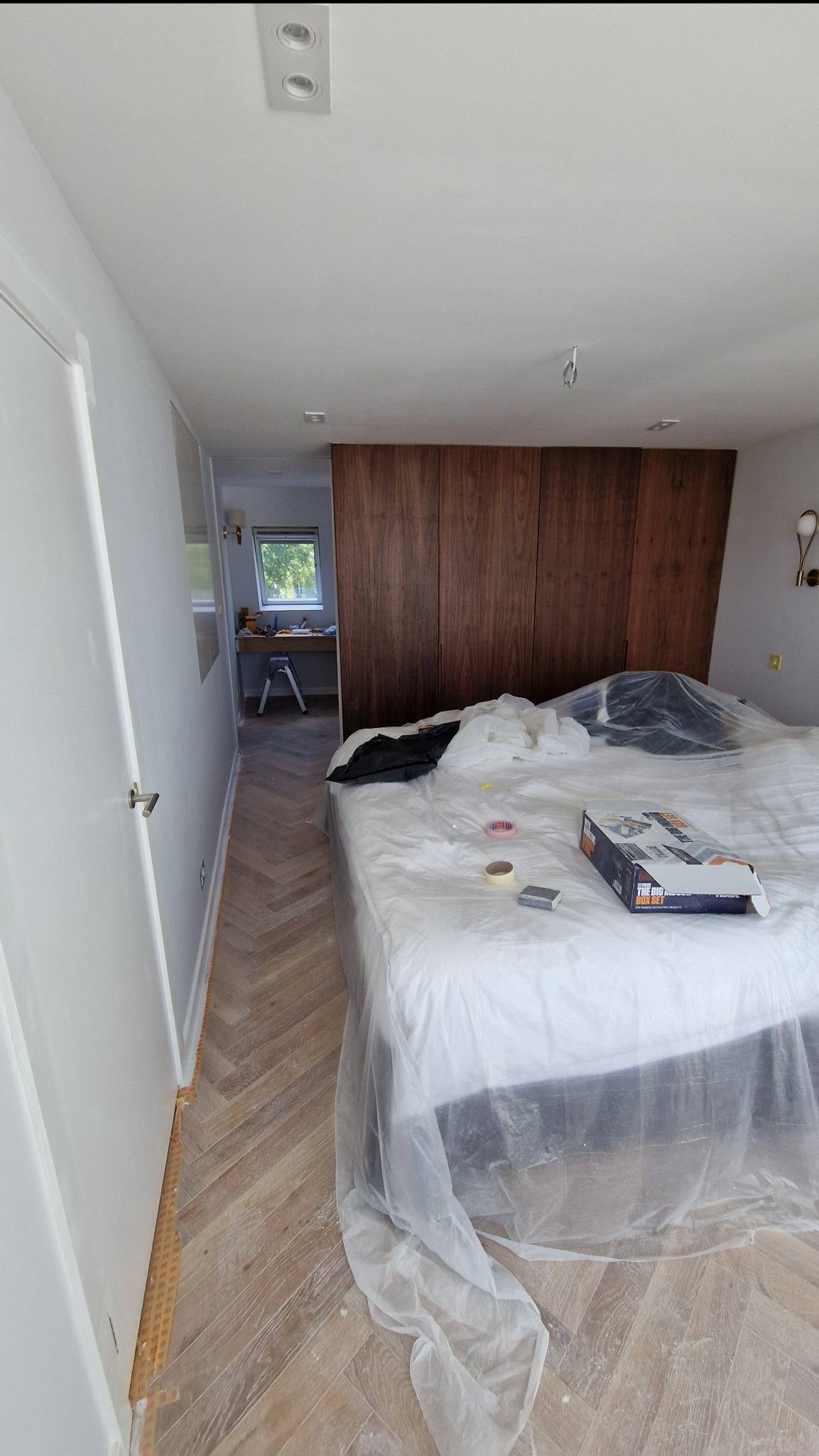 Pokój w trakcie prac wykończeniowych: zabezpieczone meble folią, widoczne narzędzia i materiały malarskie. Parkiet ułożony w jodełkę, ściany przygotowane do malowania.