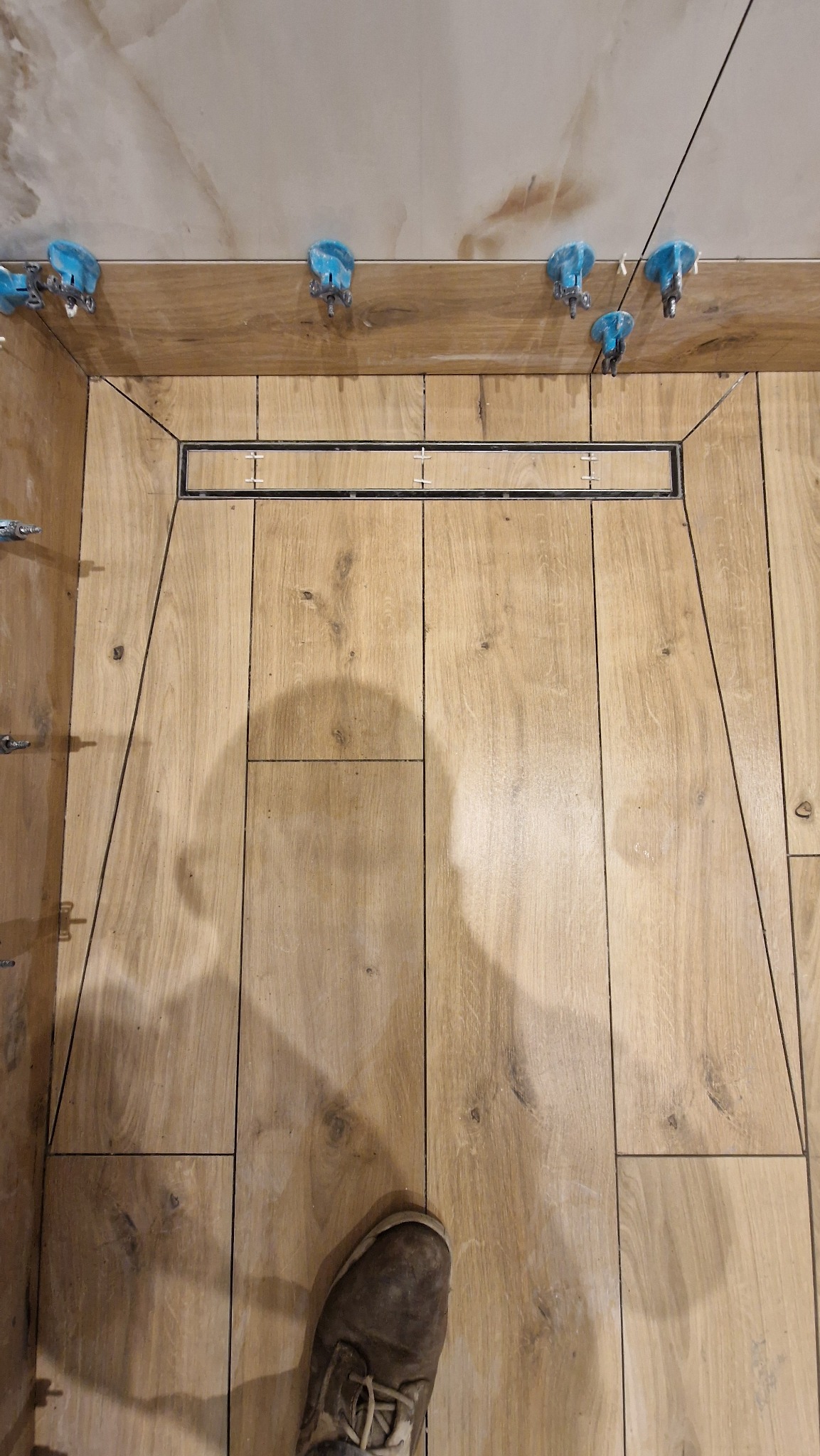 Ułożone płytki podłogowe imitujące drewno w łazience z odpływem liniowym. Widoczne niebieskie klipsy poziomujące i fragment buta.