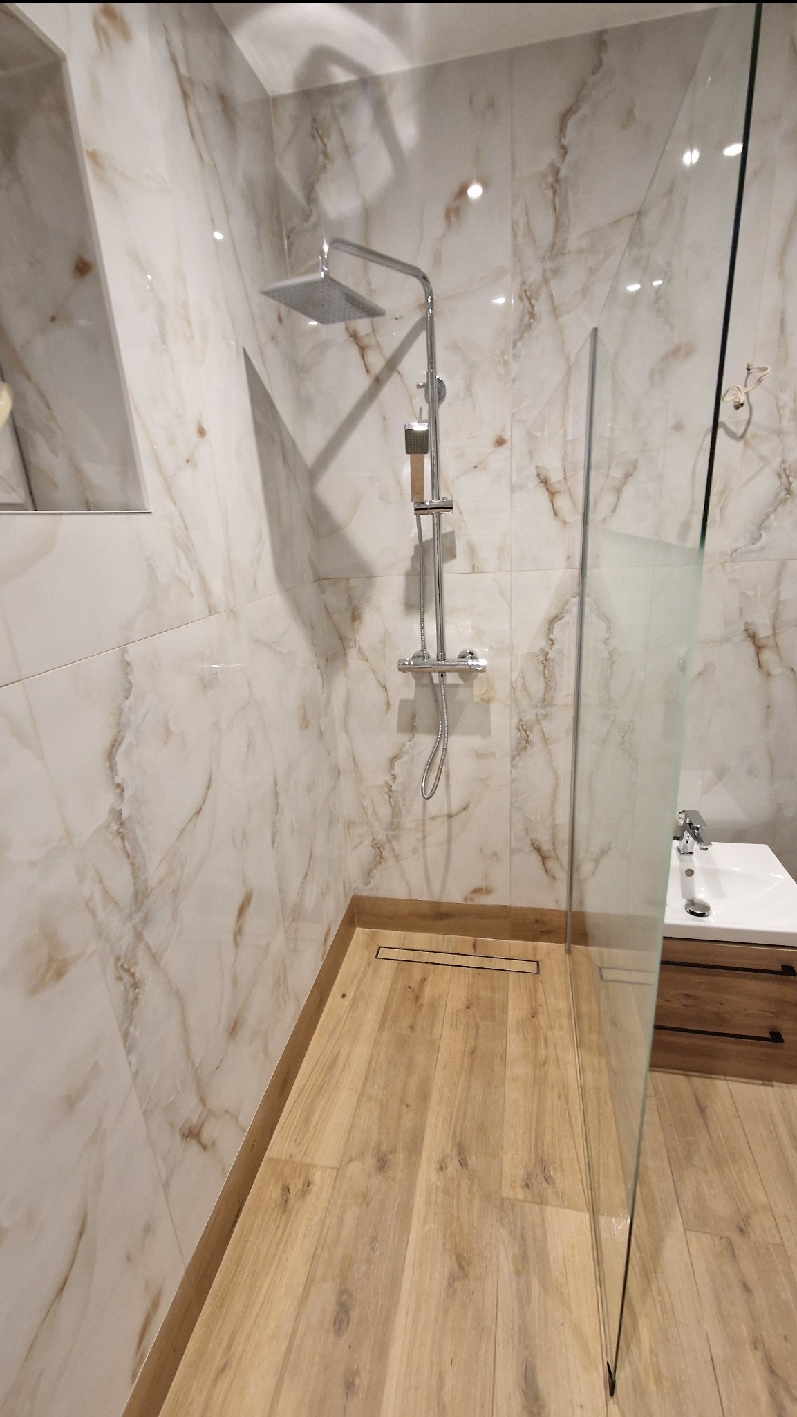 Nowoczesna łazienka z kabiną typu walk-in, wykończona płytkami imitującymi marmur i drewnopodobną podłogą. Widoczny odpływ liniowy i bateria prysznicowa.