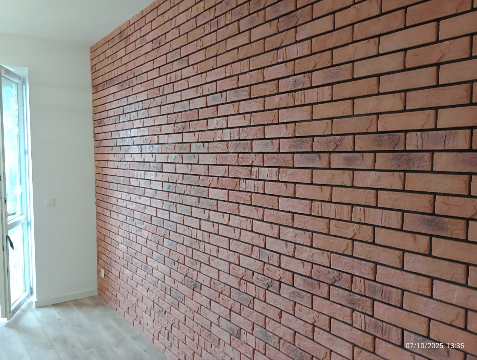 Ściana z cegły dekoracyjnej w odcieniach czerwieni, świeżo po montażu w Poniatowicach. Widoczny fragment okna i podłogi. Estetyczne wykończenie wnętrza.