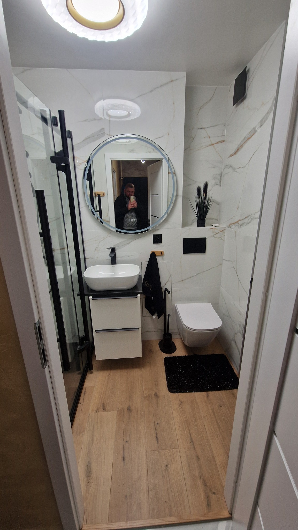 Nowoczesna łazienka z kabiną prysznicową typu walk-in, umywalką na szafce, okrągłym lustrem i drewnopodobną podłogą. Marmurowe płytki na ścianach.