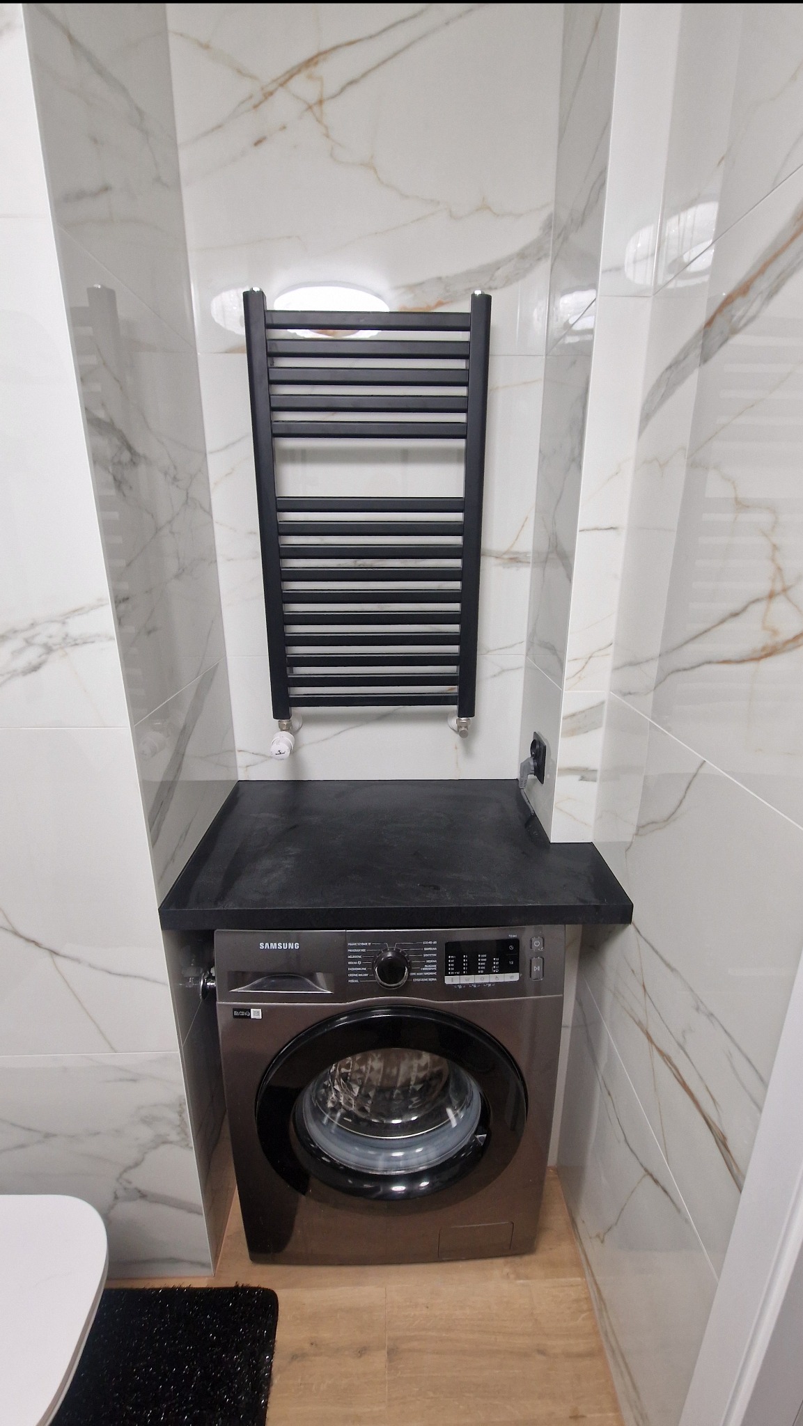 Nowoczesna łazienka z pralką Samsung pod blatem i czarnym grzejnikiem drabinkowym na tle białych płytek imitujących marmur.
