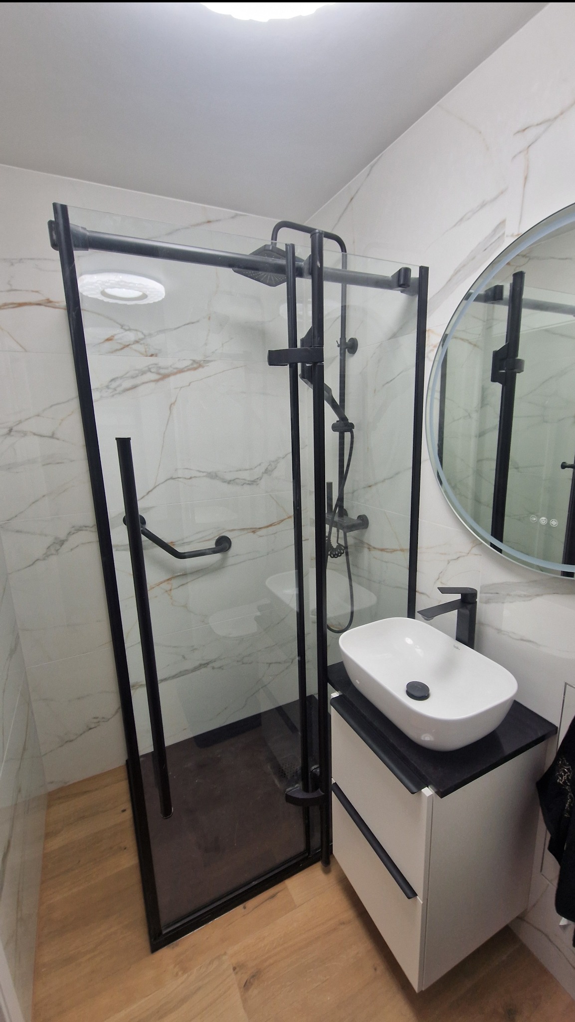 Nowoczesna łazienka z kabiną prysznicową typu walk-in z czarnymi profilami, białą umywalką nablatową i okrągłym lustrem z podświetleniem LED. Marmurowe płytki na ścianach.