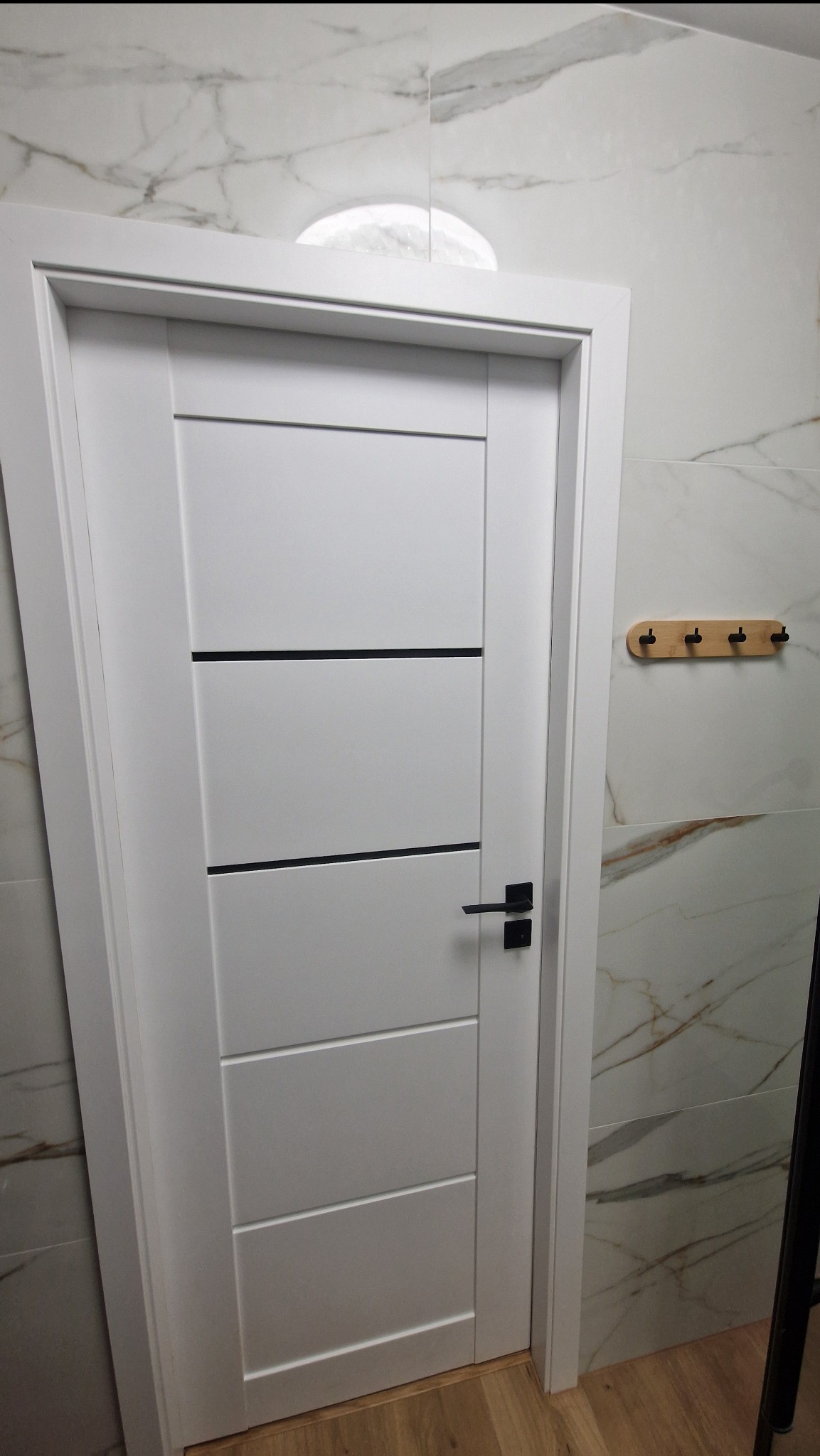 Białe drzwi z czarnymi akcentami w łazience wykończonej marmurowymi płytkami. Na ścianie drewniany wieszak z czarnymi haczykami. Widoczna podłoga z paneli.