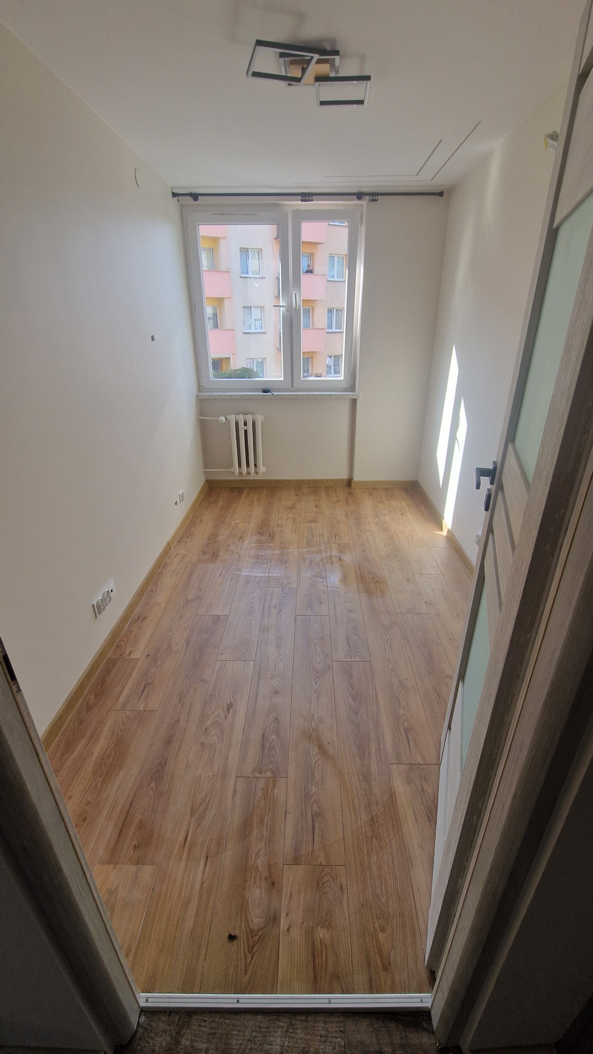 Jasne panele podłogowe w pustym pokoju z białymi ścianami i grzejnikiem pod oknem. Widok zza otwartych drzwi, nowoczesna lampa sufitowa.