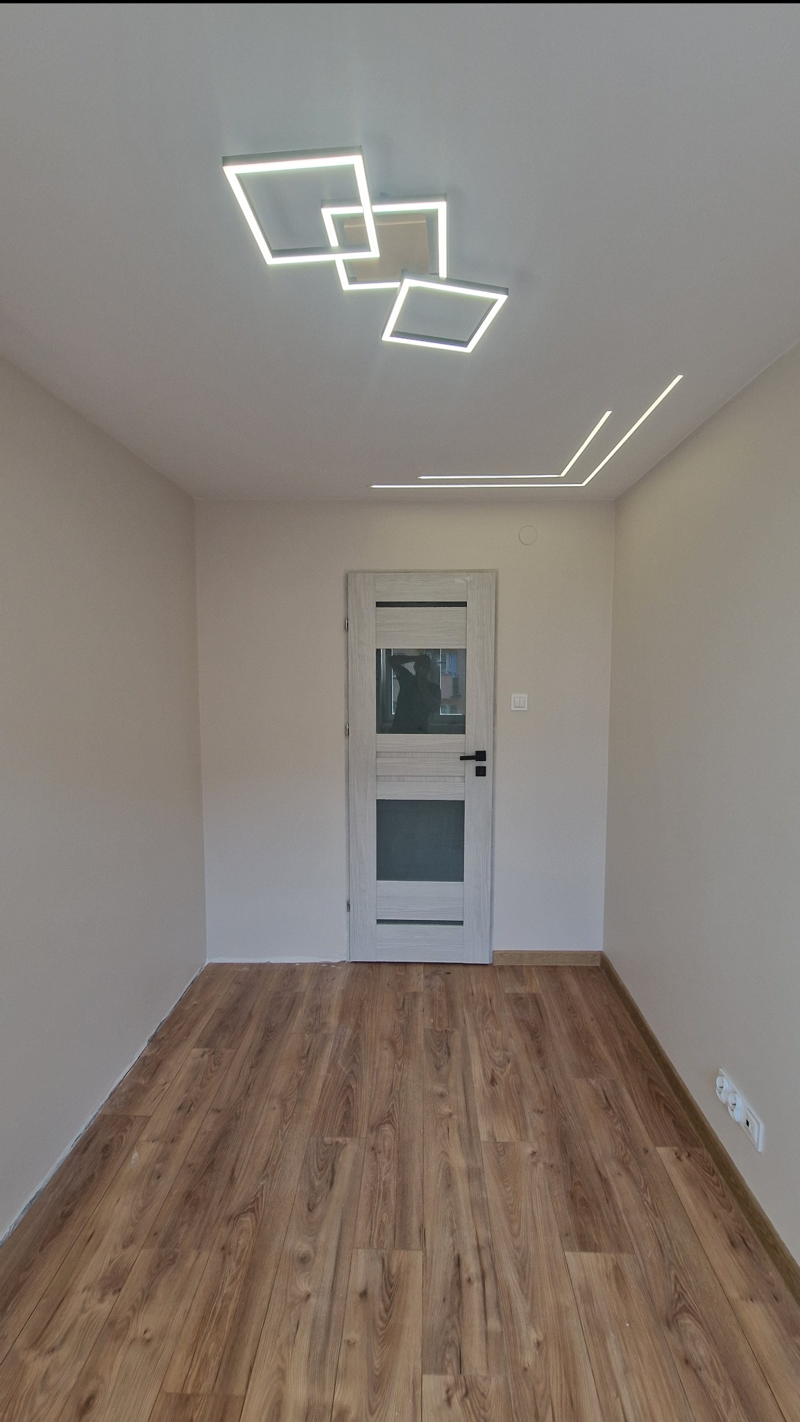 Wnętrze pokoju po remoncie: drewnopodobne panele, jasne ściany, białe drzwi z przeszkleniami i nowoczesne oświetlenie sufitowe w kształcie kwadratów i linii.