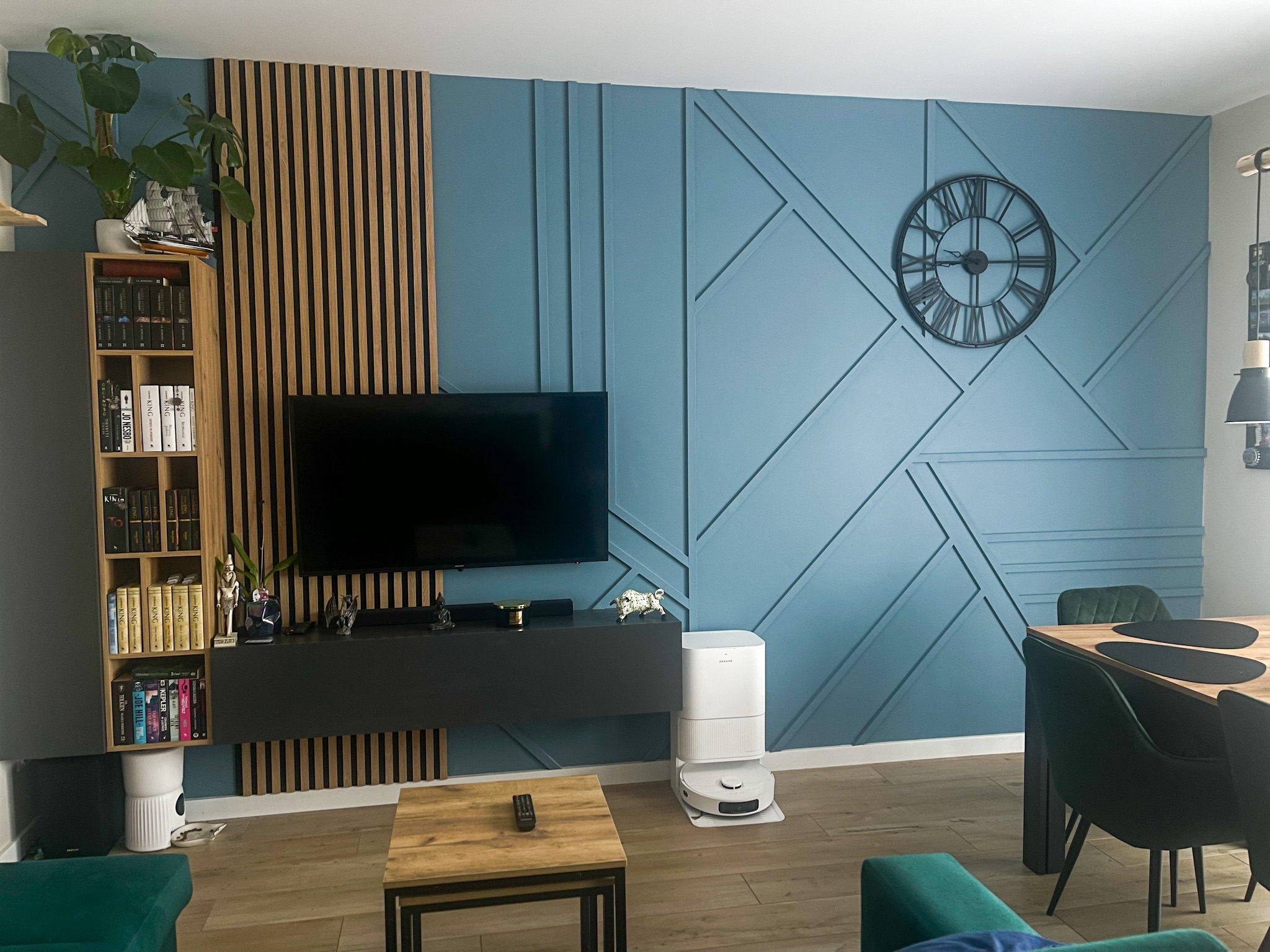 Niebieska ściana z geometrycznym wzorem i drewnianymi lamelami. Nowoczesny salon z telewizorem, regałem i robotem sprzątającym. Dekoracyjne wykończenie wnętrza.