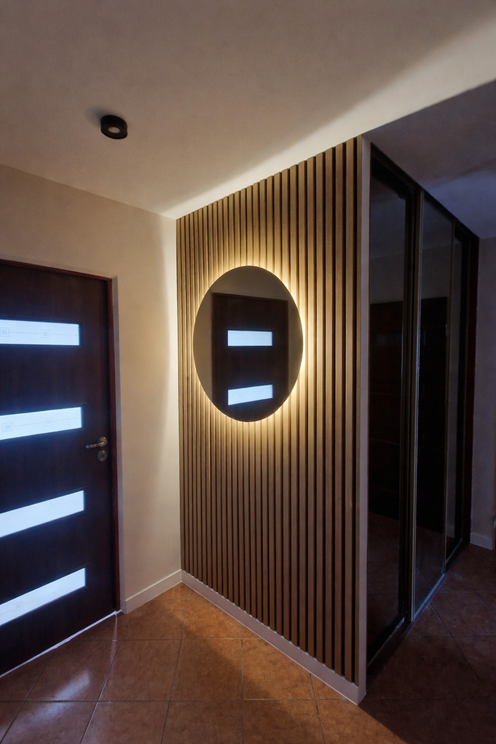 Nowoczesny przedpokój z drewnianą ścianą z lamelami i okrągłym lustrem z podświetleniem LED. Widoczna brązowa podłoga i drzwi z białymi wstawkami.