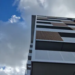 Nowoczesny budynek wielorodzinny z balkonami, ażurowe balustrady i drewniane elementy dekoracyjne, widok z dołu na tle błękitnego nieba z chmurami.