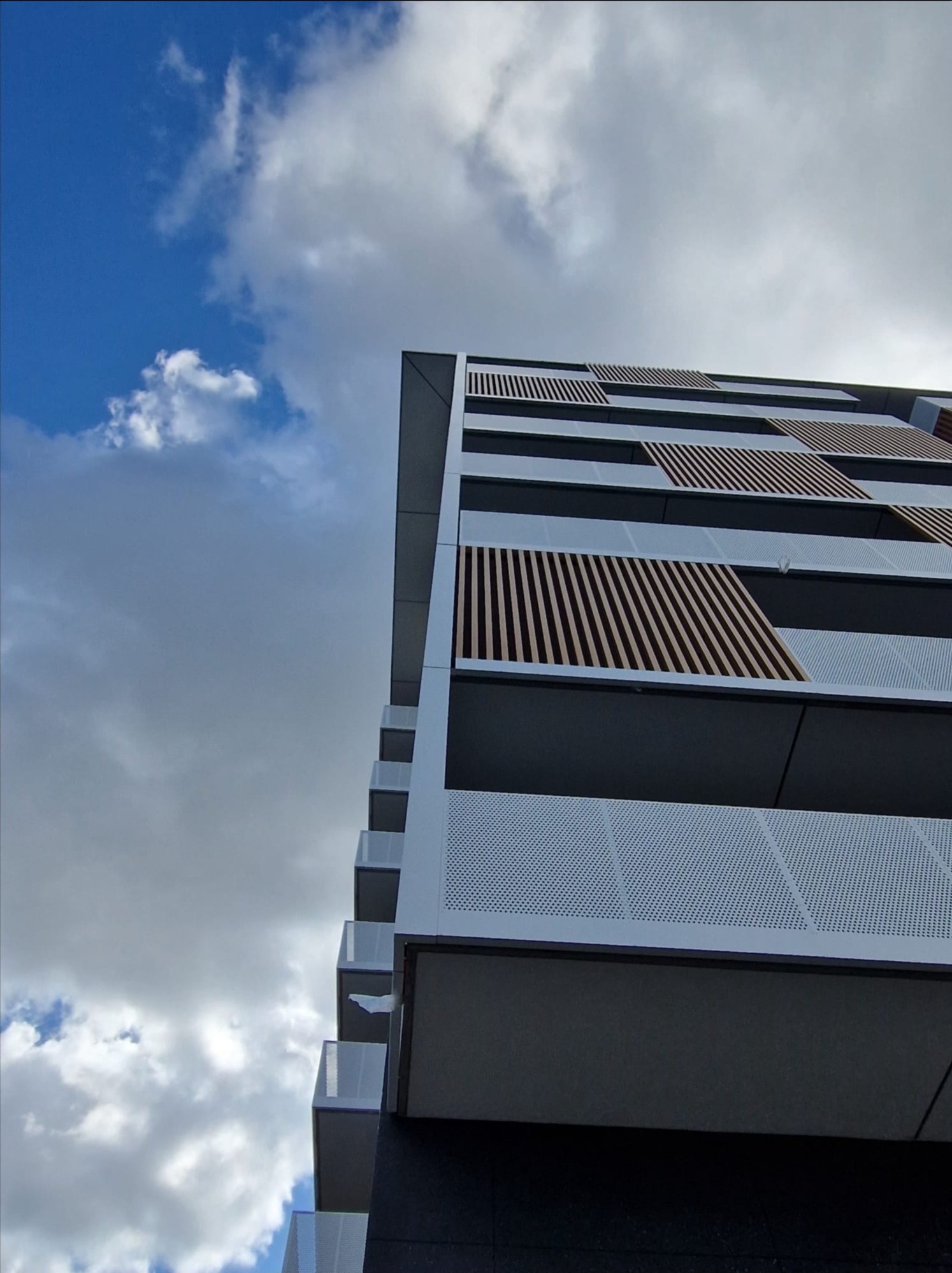 Nowoczesny budynek wielorodzinny z balkonami, ażurowe balustrady i drewniane elementy dekoracyjne, widok z dołu na tle błękitnego nieba z chmurami.