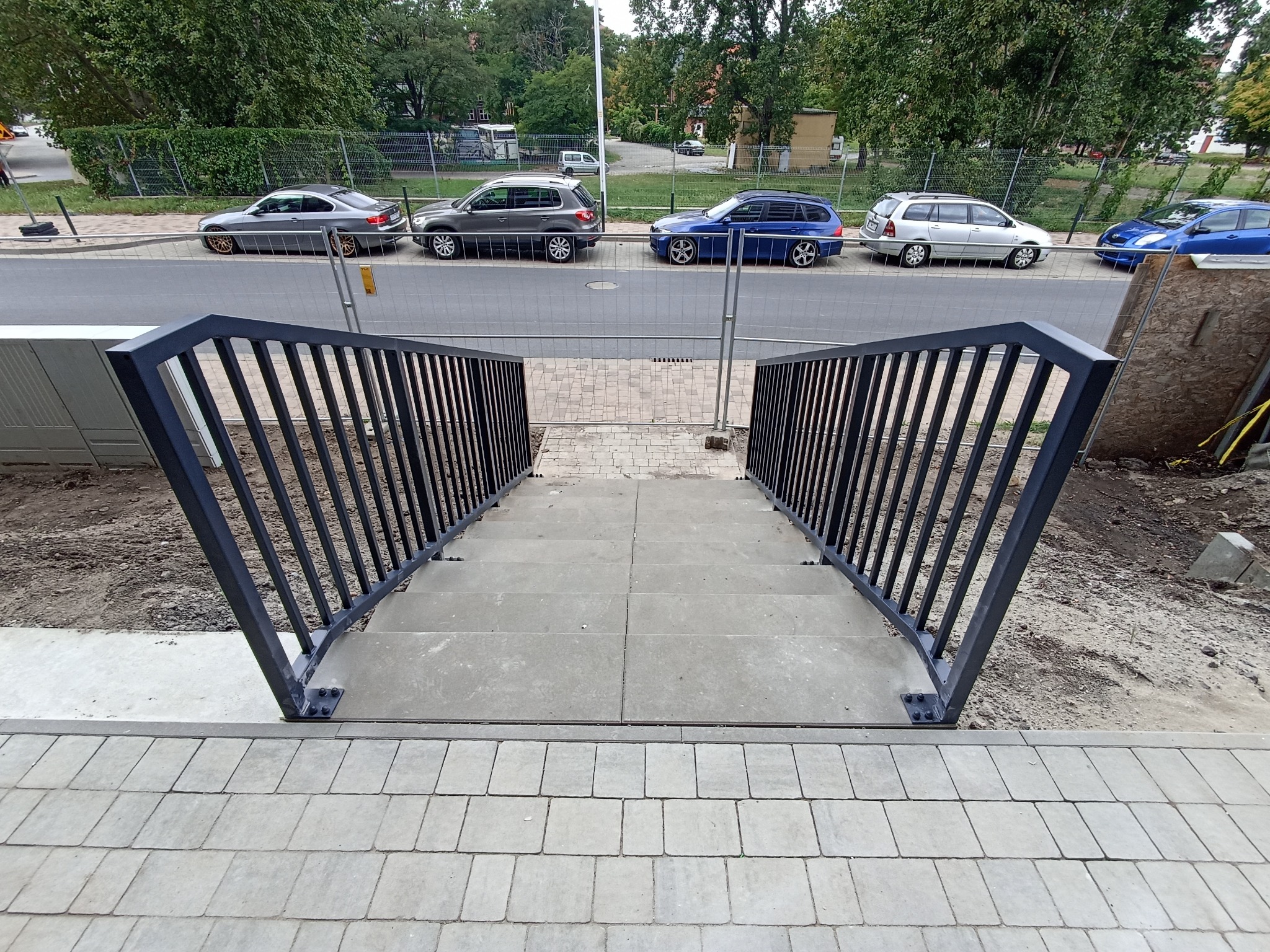 Nowoczesne schody zewnętrzne z ciemną metalową poręczą, widok z góry. Ułożona kostka brukowa i samochody w tle.