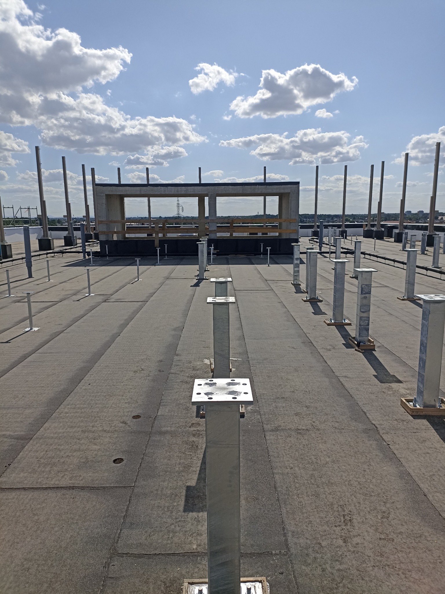 Metalowe słupki montażowe na dachu budynku, przygotowane do instalacji konstrukcji. Widoczne mocowania i elementy montażowe. W tle betonowy szkielet budynku i błękitne niebo.