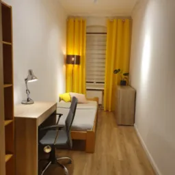 Pokój młodzieżowy z drewnianym biurkiem, krzesłem obrotowym i łóżkiem. Żółte zasłony w oknie. Na podłodze panele. Oświetlenie sufitowe punktowe.