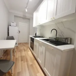 Jasna kuchnia z białymi frontami szafek z frezowanymi panelami, złote uchwyty, marmurowy backsplash i drewniana podłoga. Oświetlenie szynowe.