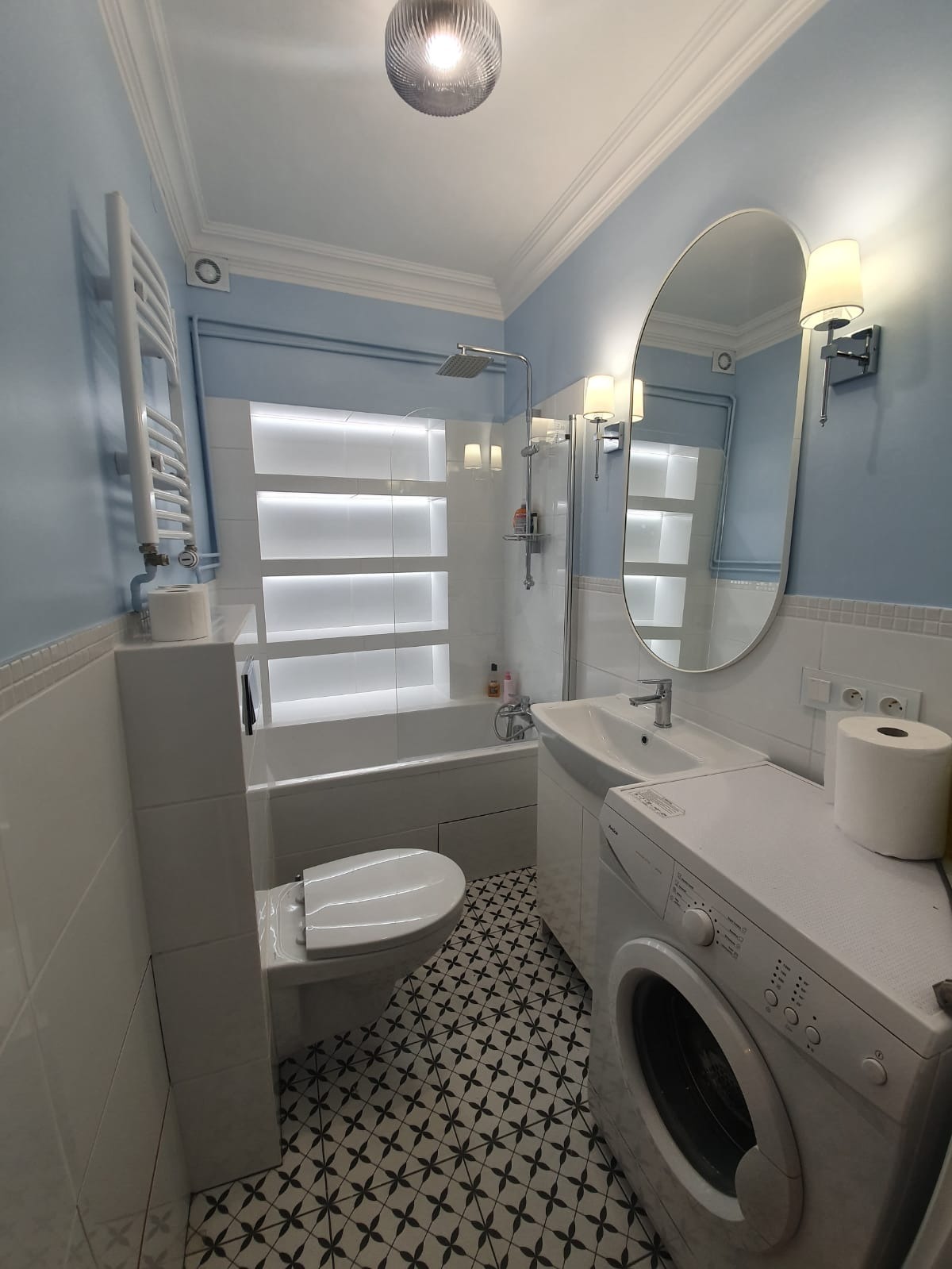 Wykończona łazienka w bieli i błękicie z wanną, umywalką, toaletą i pralką. Podłoga w czarno-białe wzory. Oświetlenie sufitowe i ścienne.