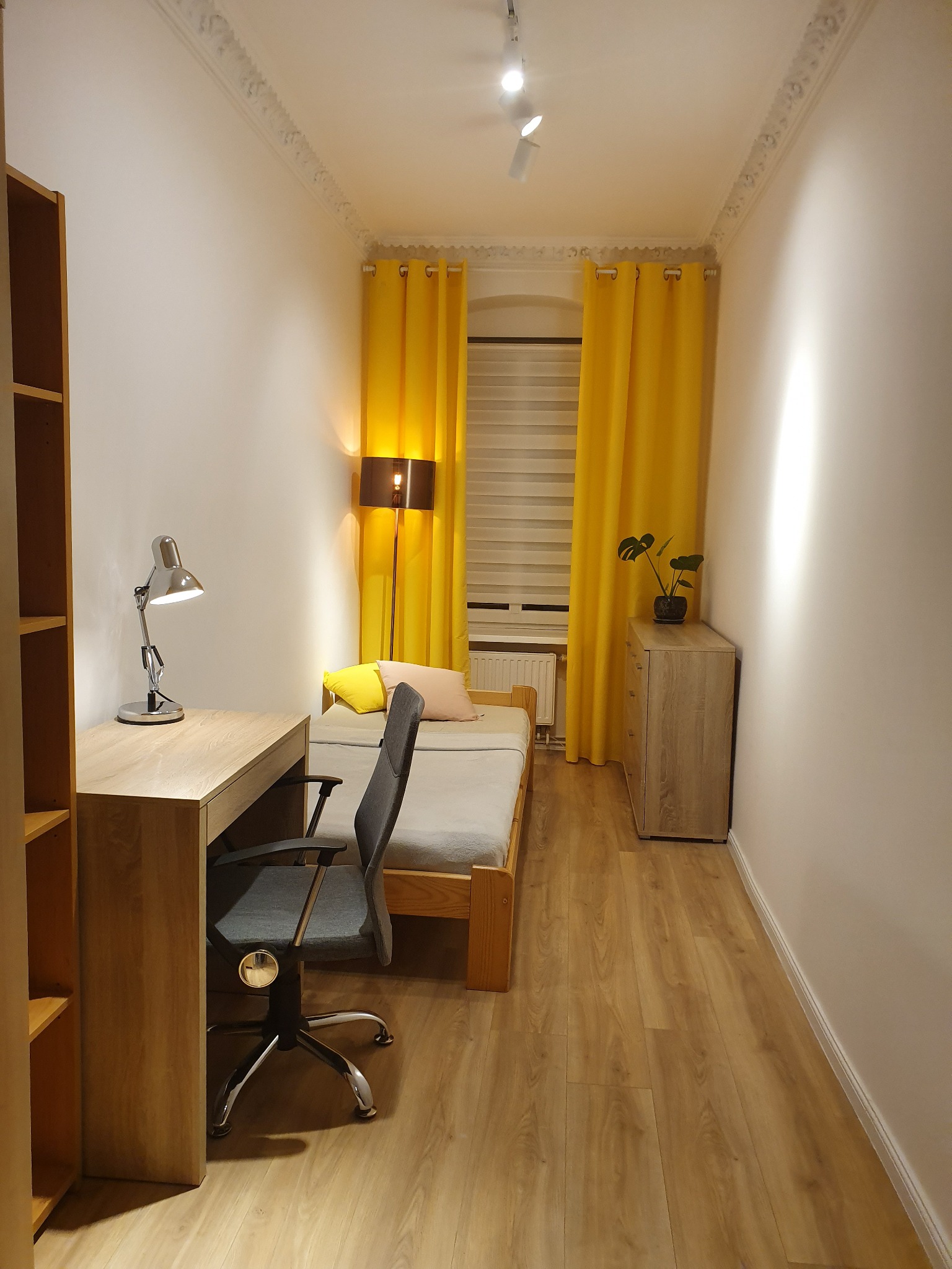 Pokój młodzieżowy z drewnianym biurkiem, krzesłem obrotowym i łóżkiem. Żółte zasłony w oknie. Na podłodze panele. Oświetlenie sufitowe punktowe.