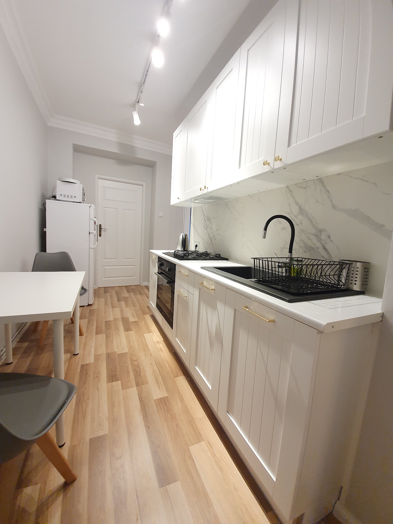 Jasna kuchnia z białymi frontami szafek z frezowanymi panelami, złote uchwyty, marmurowy backsplash i drewniana podłoga. Oświetlenie szynowe.