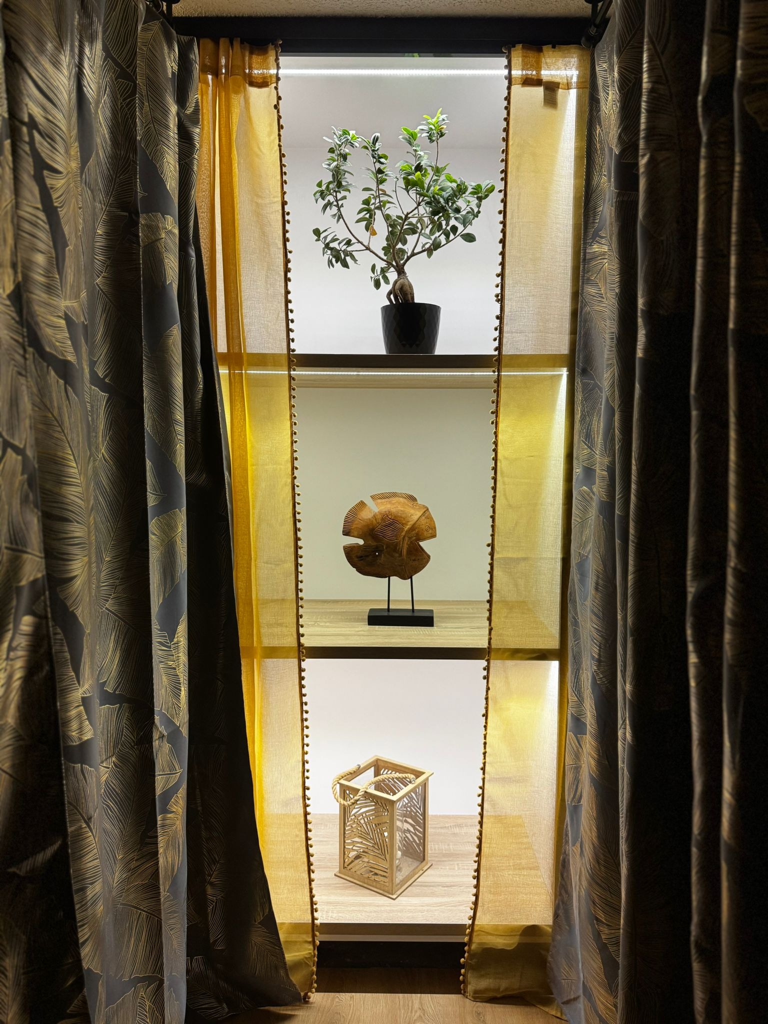 Dekoracyjna, podświetlana półka z rośliną, drewnianą rzeźbą i lampionem, przesłonięta zasłonami w odcieniach szarości i złota, tworząca przytulny klimat.