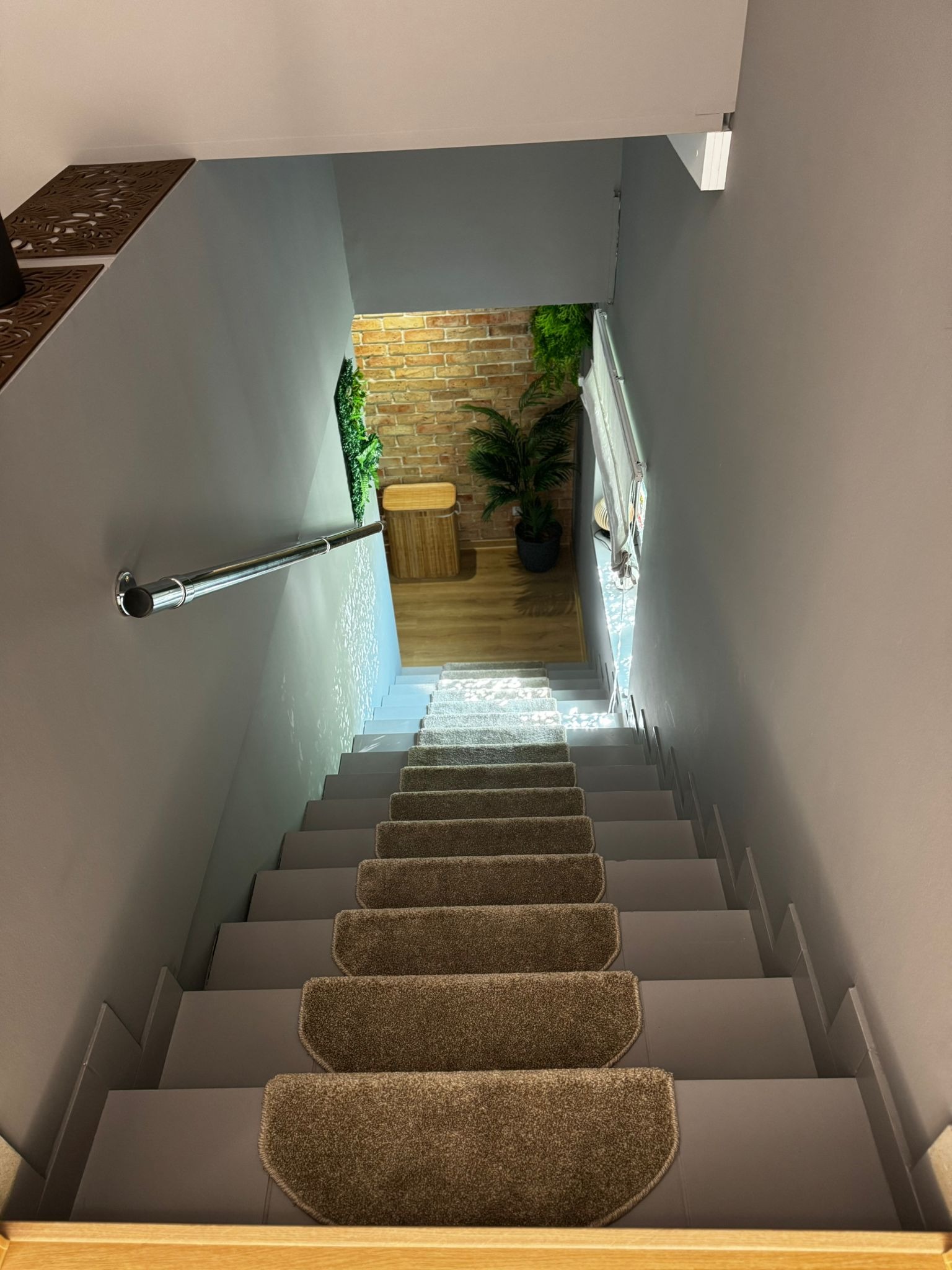 Schody w dół z szarymi stopniami i brązowymi nakładkami dywanowymi, widok z góry na jasne, urządzone pomieszczenie na końcu biegu.