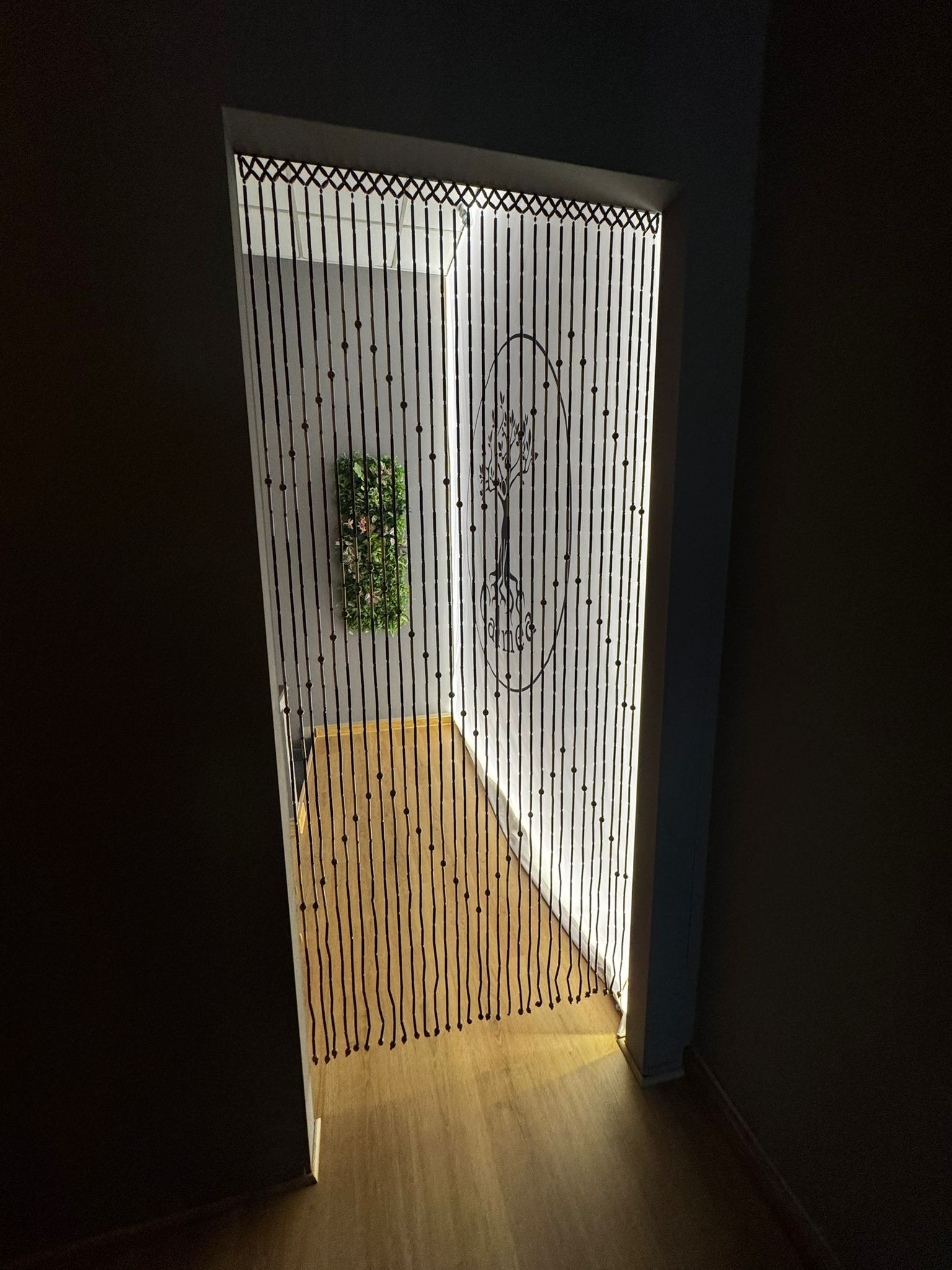 Wejście do pokoju z dekoracyjną zasłoną z koralików i motywem drzewa. W tle ściana z zieloną dekoracją roślinną. Ciepłe oświetlenie tworzy przytulną atmosferę.