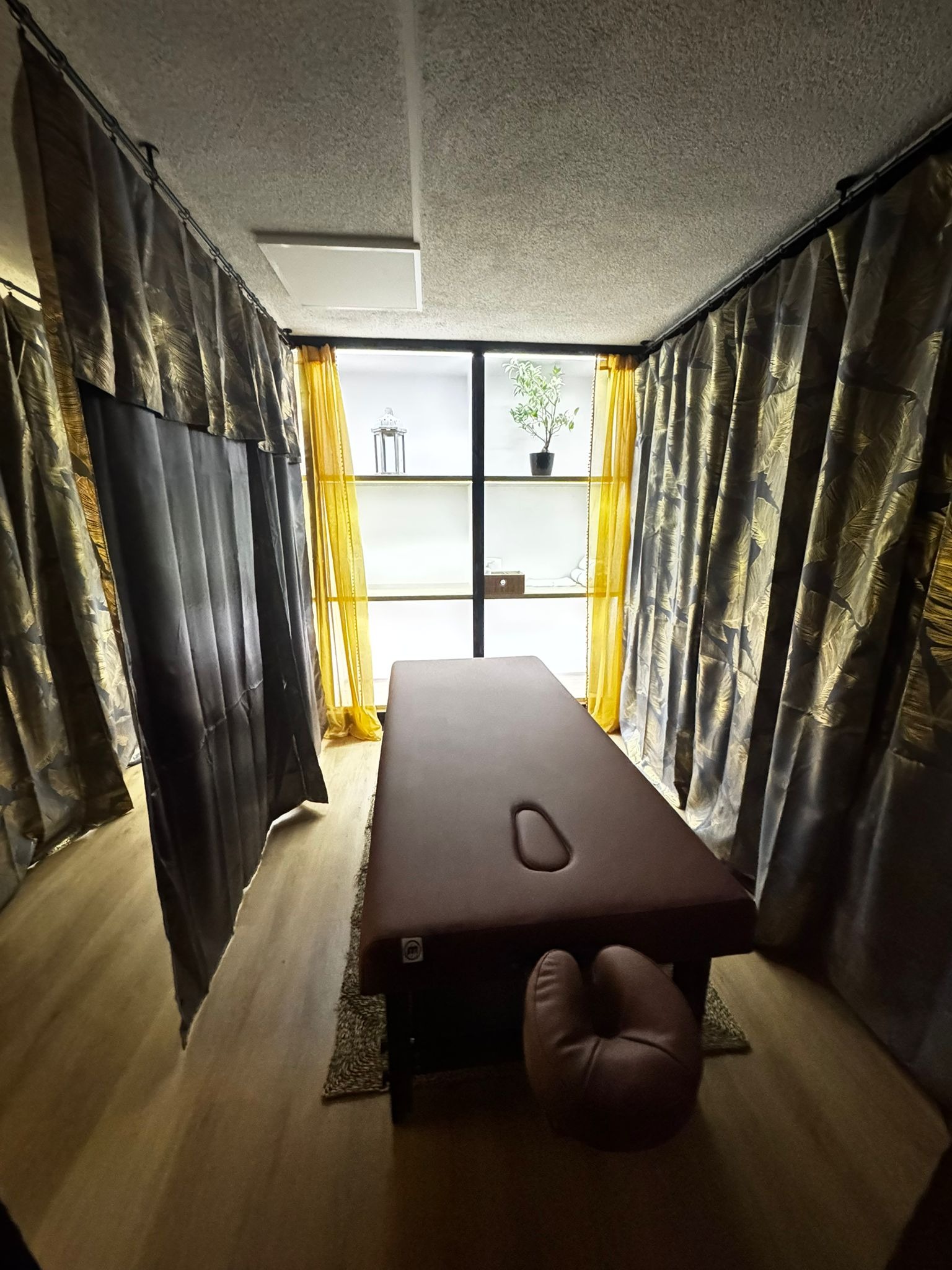 Brązowy stół do masażu z otworem na twarz, otoczony zasłonami w salonie z półkami i roślinami. Przytulna aranżacja wnętrza, ciepłe oświetlenie.