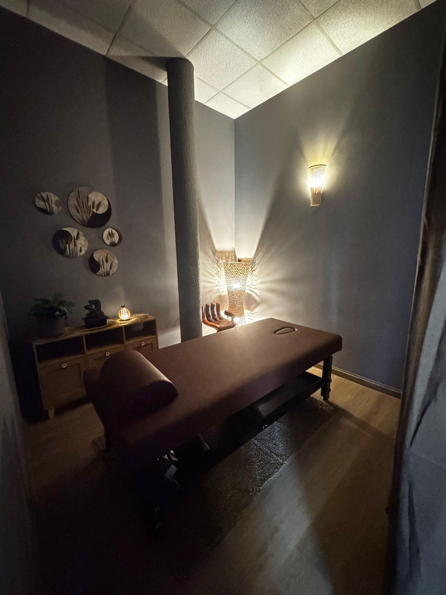 Ciemny gabinet z brązowym łóżkiem do masażu, oświetlony lampką nocną i kinkietem. Dekoracyjne talerze na ścianie i szafka z rośliną dodają przytulności wnętrzu.