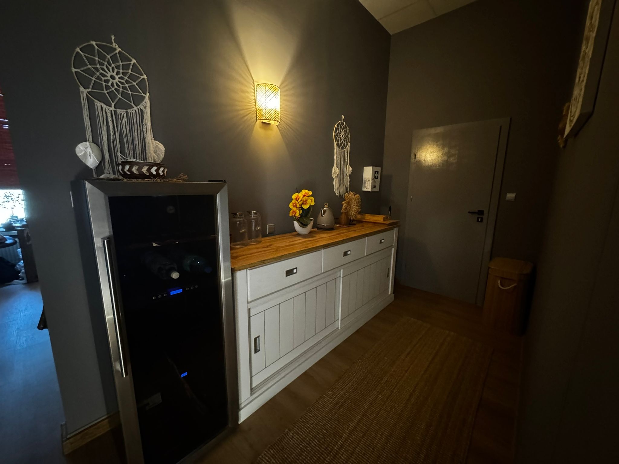 Wnętrze gabinetu z nastrojowym oświetleniem, dekoracjami typu łapacz snów i komodą z drewnianym blatem oraz lodówką na wino. Przytulny klimat.