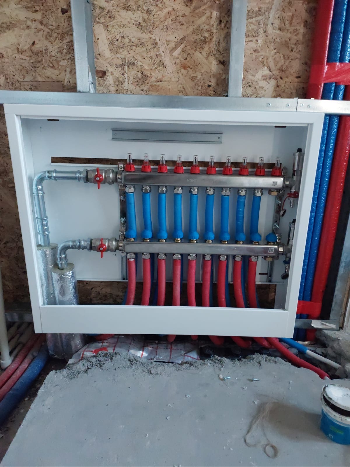 Rozdzielacz ogrzewania podłogowego w białej obudowie, z widocznymi czerwonymi i niebieskimi rurami, zamontowany na ścianie z płyty OSB. Widoczne fragmenty instalacji w podłodze.