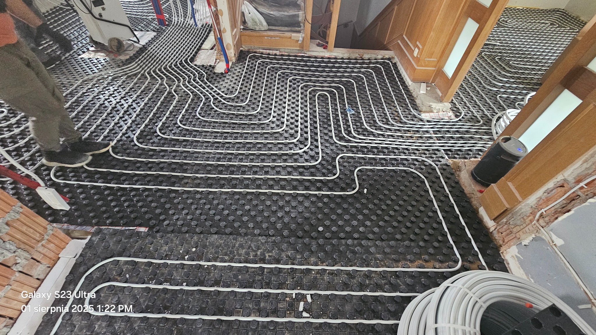 Układanie białych rur ogrzewania podłogowego na czarnej macie izolacyjnej w pomieszczeniu w trakcie remontu. Widoczne fragmenty ścian i rur.