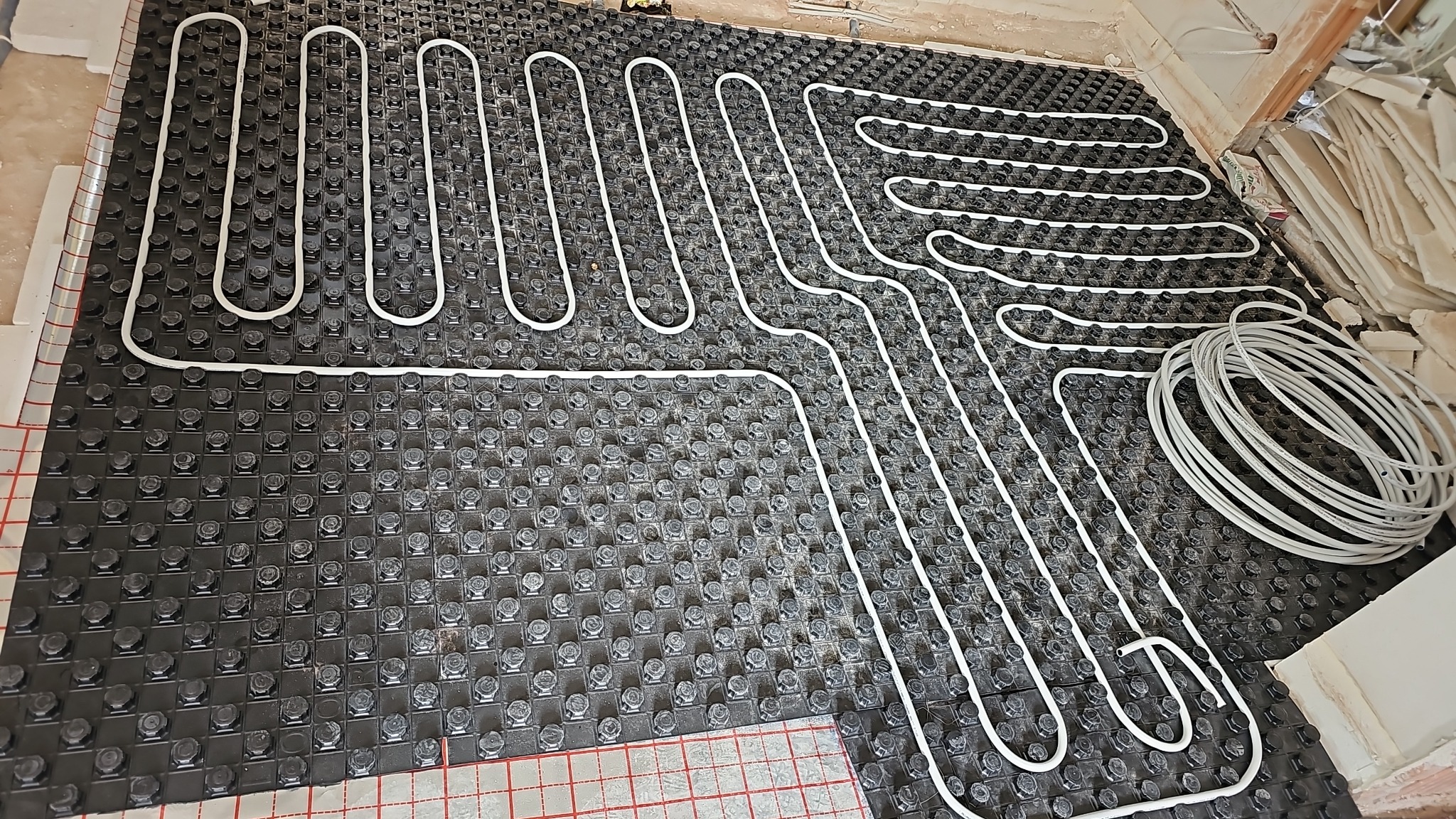 Białe rurki ogrzewania podłogowego ułożone na czarnej macie z wypustkami, widoczne fragmenty posadzki i zwój rur.