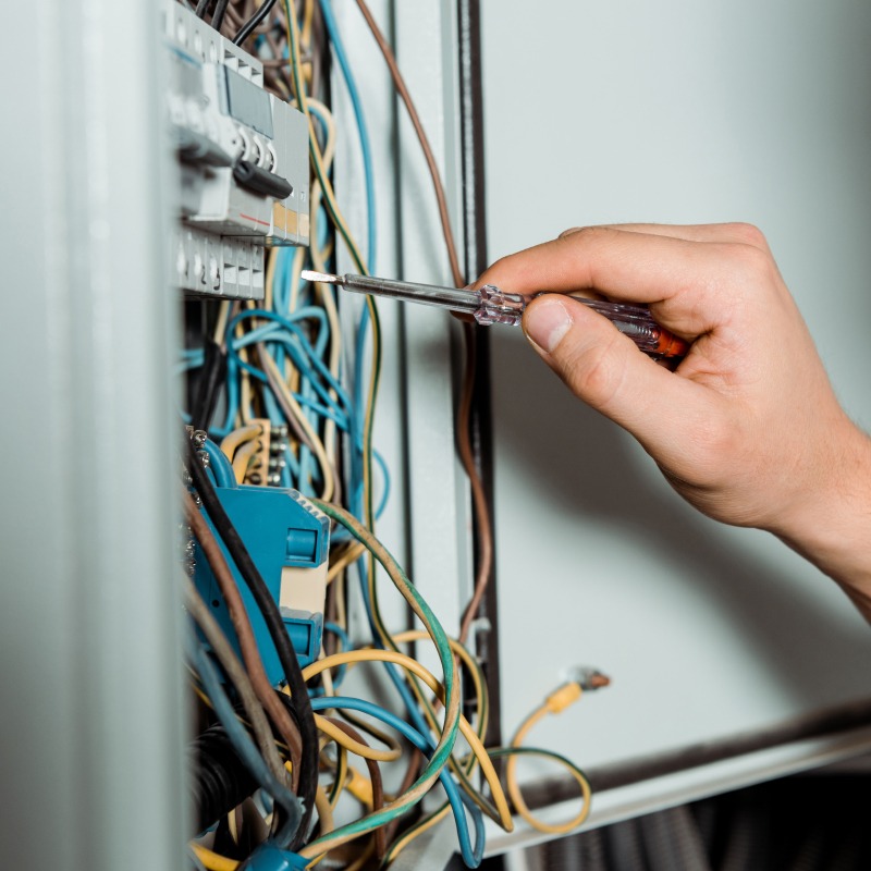 Elektryk testuje napięcie w skrzynce bezpiecznikowej za pomocą próbnika napięcia. Widoczne kolorowe przewody i bezpieczniki.