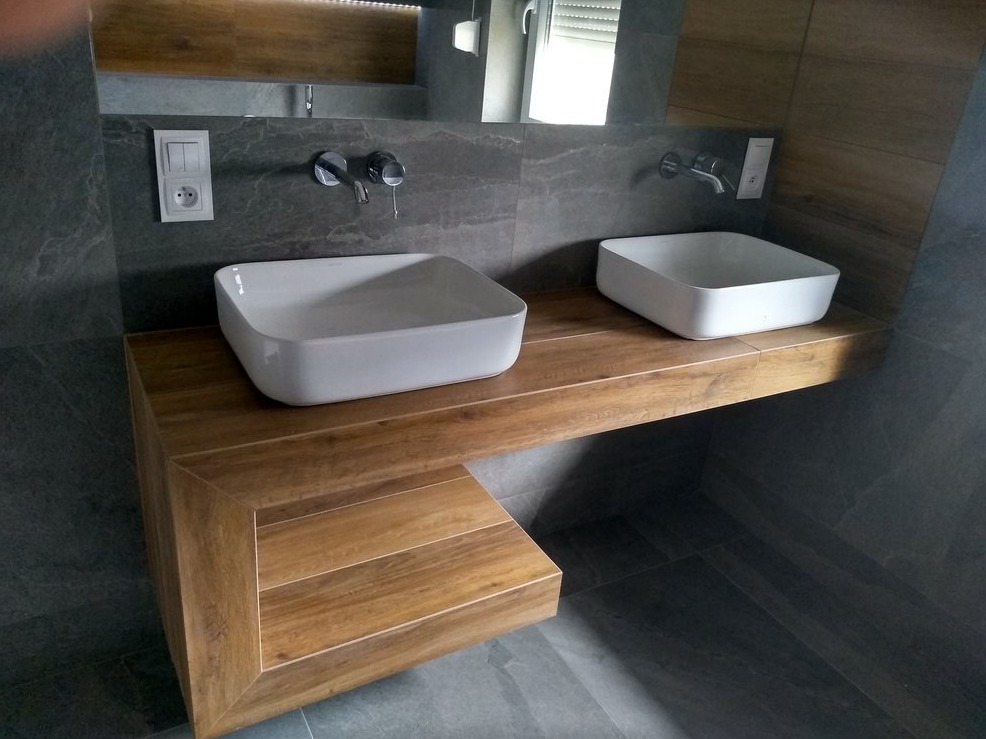 Nowoczesna łazienka: dwie białe umywalki nablatowe na drewnianym blacie z płytek, szare płytki na ścianach i podłodze, chromowana armatura.