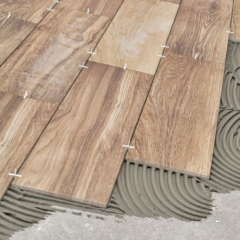 Układanie płytek imitujących drewno na podłodze, widoczny klej i krzyżyki dystansowe. Ujęcie z góry, jasne oświetlenie.