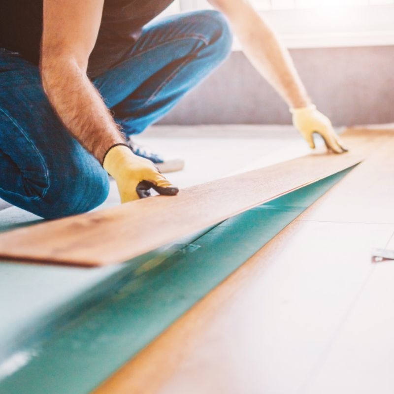 Montaż paneli podłogowych: mężczyzna w rękawicach układa drewniane panele na zielonej podkładce, widoczne dłonie i fragment podłogi w jasnym pomieszczeniu.