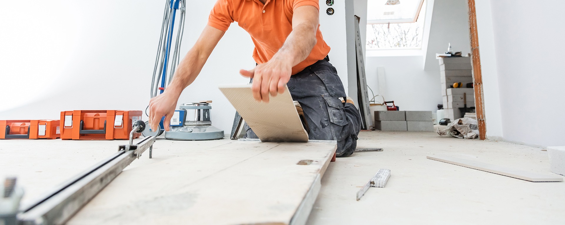 Glazurnik docina beżową płytkę ceramiczną na profesjonalnej maszynce. W tle widoczne narzędzia, pojemniki i materiały budowlane. Ujęcie w trakcie pracy.