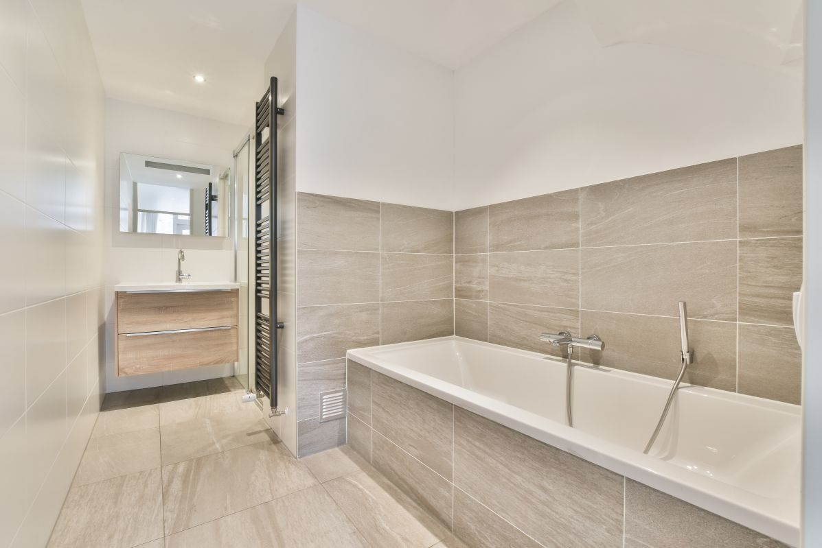 Jasna łazienka z beżowymi płytkami, białą wanną i drewnianą szafką pod umywalką. Grzejnik drabinkowy obok wanny. Minimalistyczny design.