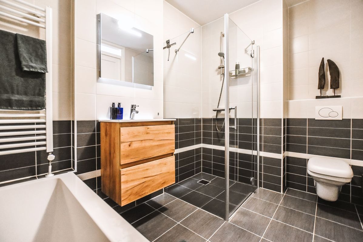 Nowoczesna łazienka z kabiną prysznicową, drewnianą szafką pod umywalką i czarno-białymi płytkami. Grzejnik drabinkowy z ręcznikiem.