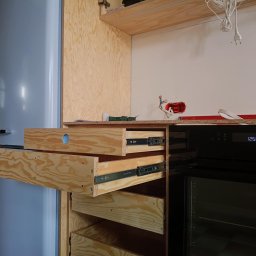 .bygga - USŁUGI MICHAŁ BIGUS - Wnętrze kuchni w trakcie montażu: drewniane szuflady z widocznymi prowadnicami, zabudowa lodówki w odcieniach błękitu i sprzęt AGD. Widoczne elementy instalacji elektrycznej.