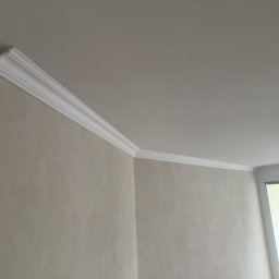 .bygga - USŁUGI MICHAŁ BIGUS - Wnętrze po montażu białej listwy dekoracyjnej na styku ściany i sufitu. Jasne ściany z delikatną fakturą. Fragment drzwi w tle. Ujęcie z dołu, podkreślające detale wykończenia.