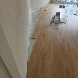 .bygga - USŁUGI MICHAŁ BIGUS - Układanie podłogi drewnianej w trakcie prac. Widoczne panele, podkład, narzędzia i fragment ściany. Jasne wnętrze, etap montażu.