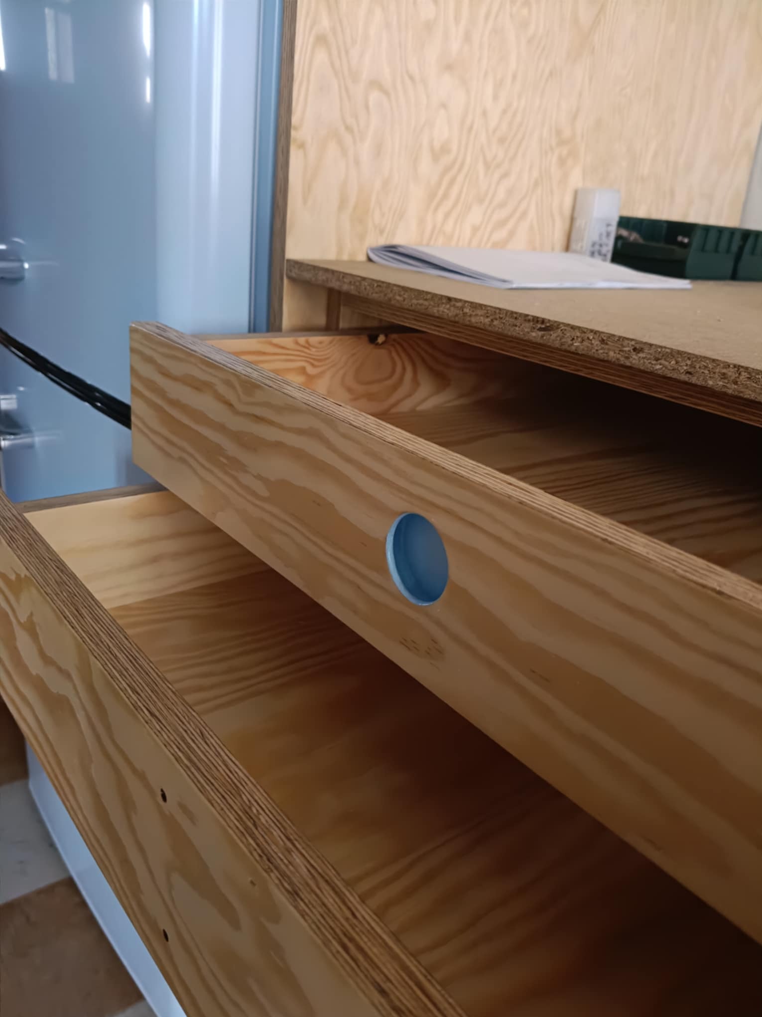 Otwarta drewniana szuflada z wywierconym otworem, widoczna struktura drewna i fragment niebieskiej obudowy. Szuflada wysunięta, wnętrze puste.