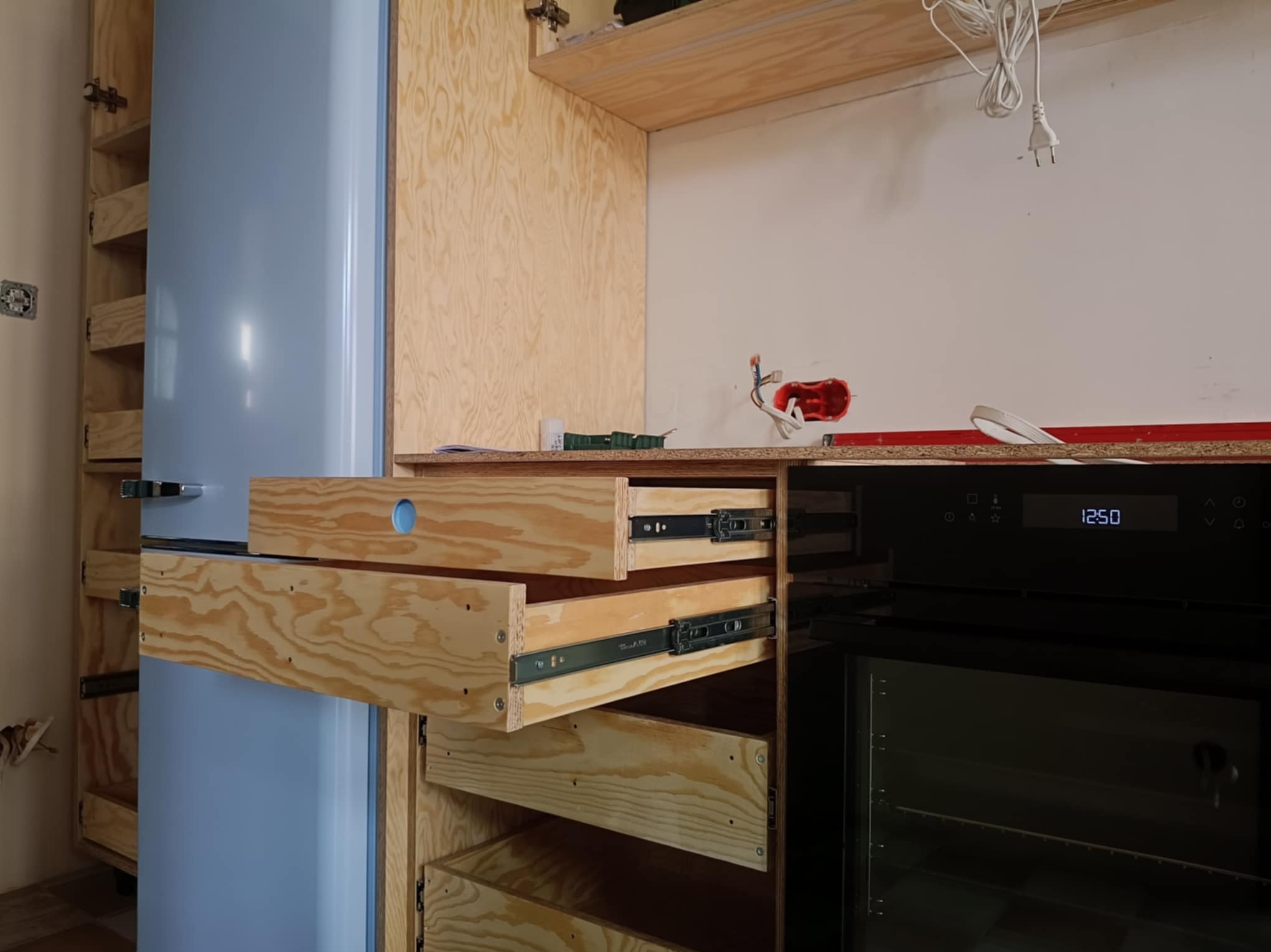 Wnętrze kuchni w trakcie montażu: drewniane szuflady z widocznymi prowadnicami, zabudowa lodówki w odcieniach błękitu i sprzęt AGD. Widoczne elementy instalacji elektrycznej.