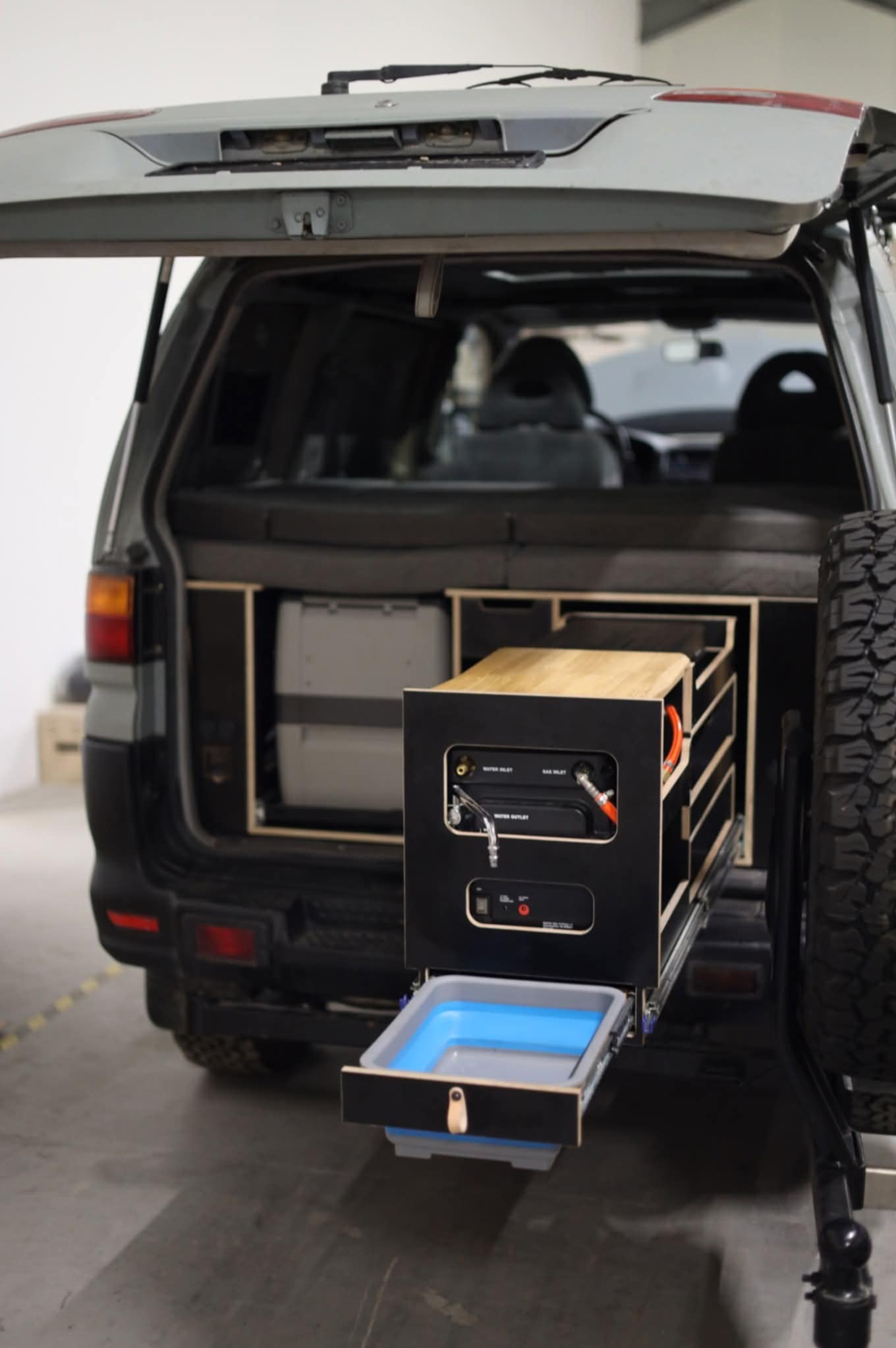Zabudowa bagażnika auta z wysuwanymi modułami: szuflada z umywalką, zasilanie i lodówka. Widoczny zapasowy koło i wnętrze pojazdu.