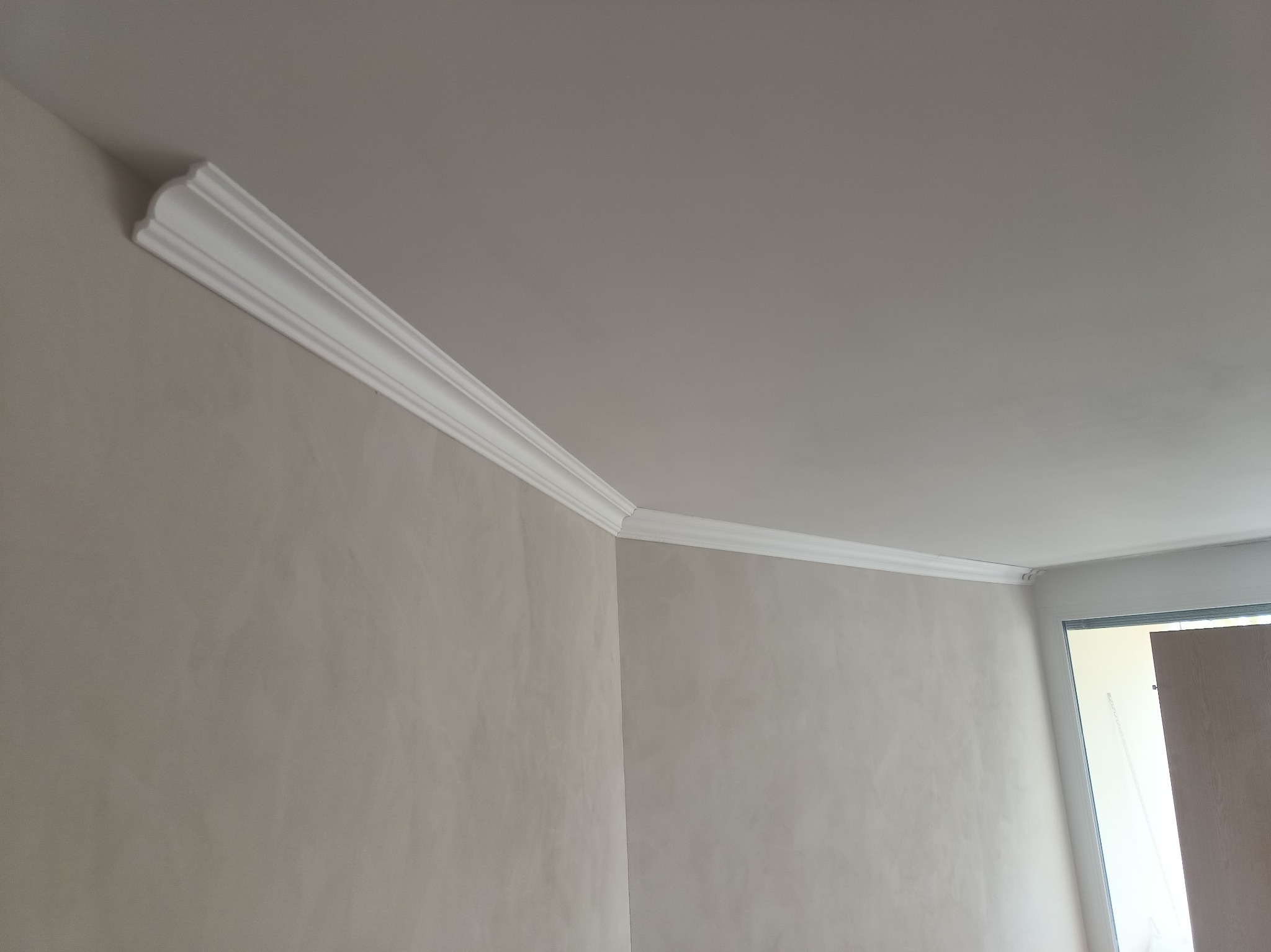 Wnętrze po montażu białej listwy dekoracyjnej na styku ściany i sufitu. Jasne ściany z delikatną fakturą. Fragment drzwi w tle. Ujęcie z dołu, podkreślające detale wykończenia.