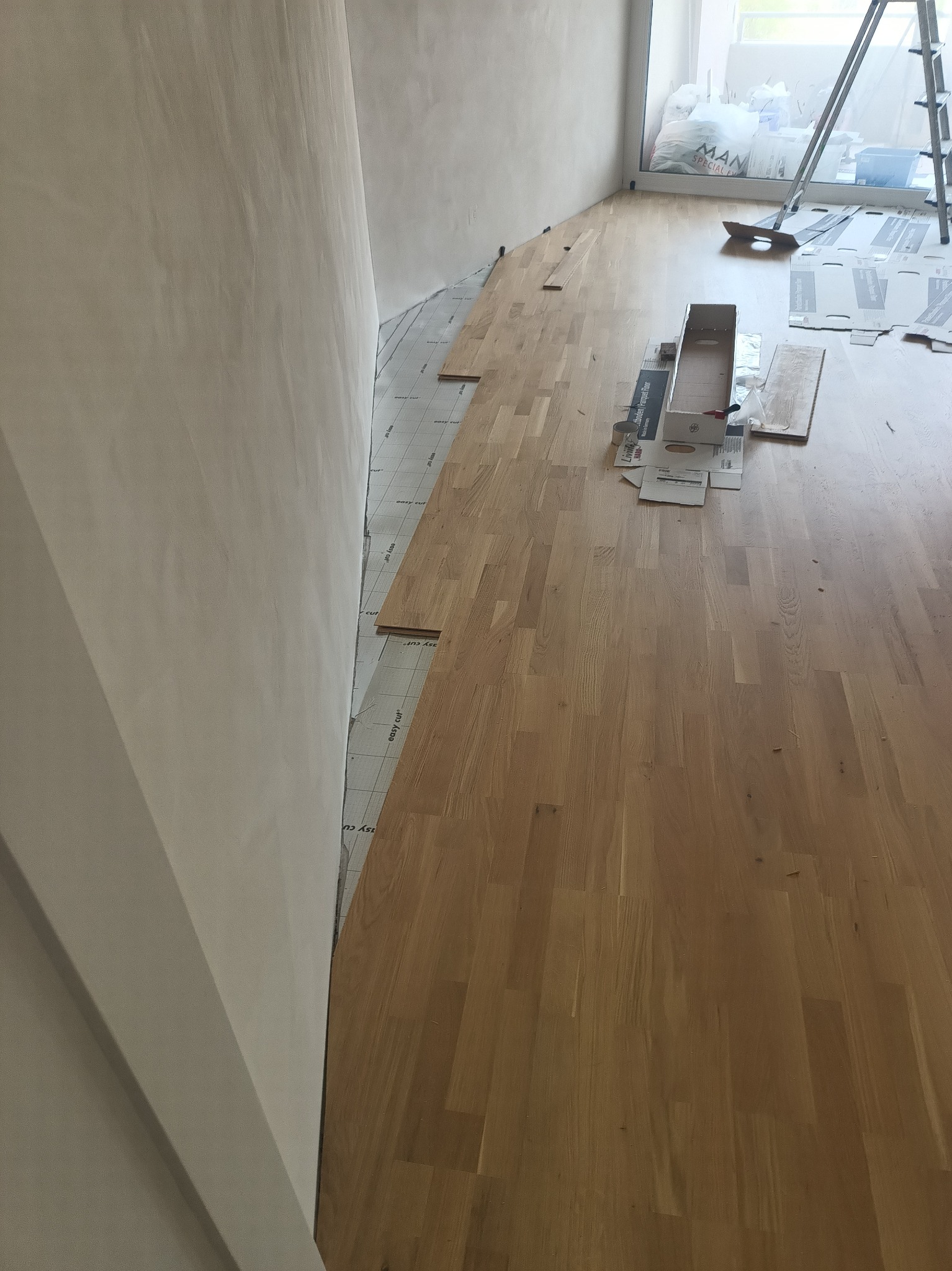 Układanie podłogi drewnianej w trakcie prac. Widoczne panele, podkład, narzędzia i fragment ściany. Jasne wnętrze, etap montażu.